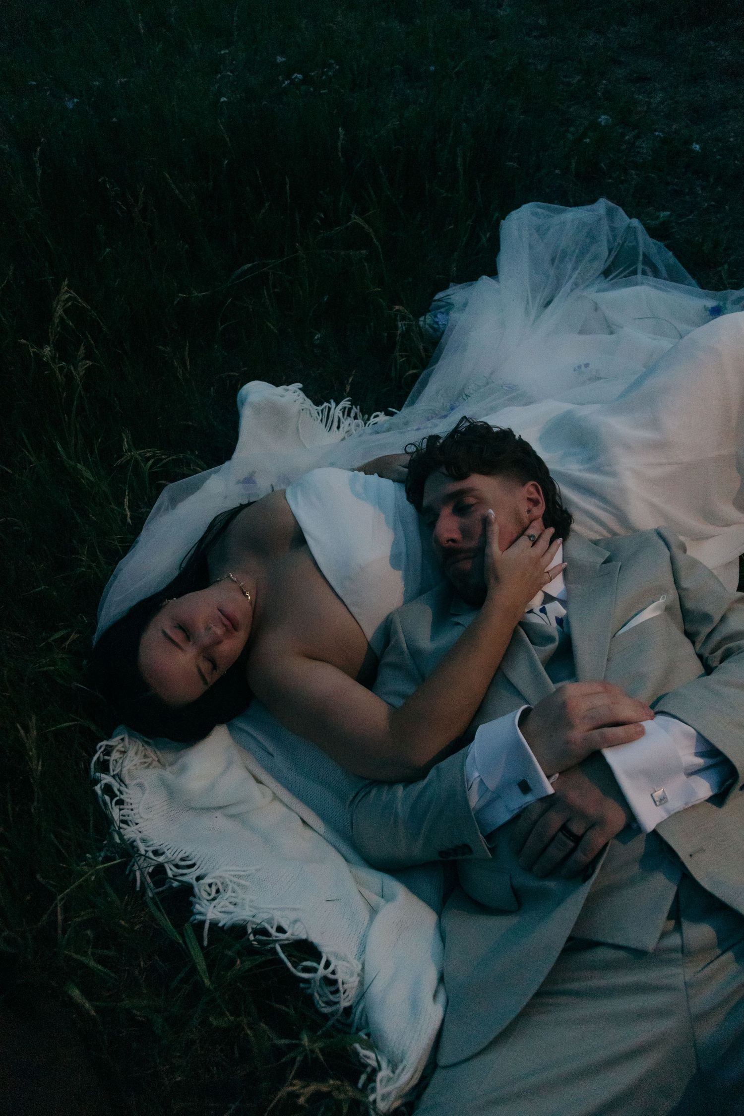 Bride and groom lying on grass at dusk, she wears white dress with veil draped over them both.
