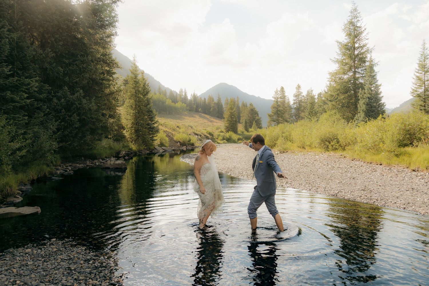 Bride and groom wading through a mountain stream in wedding attire, surrounded by pine trees and scenic alpine landscape.
