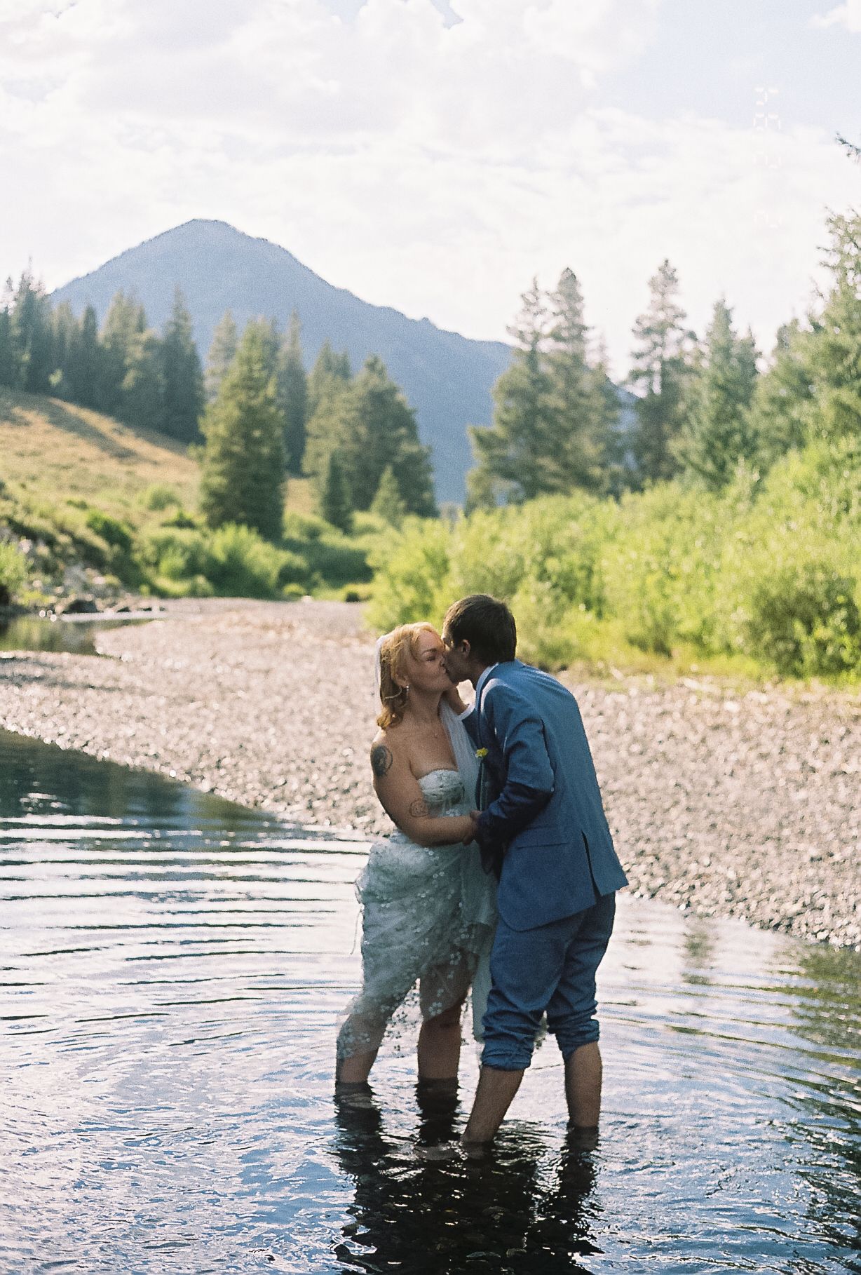 Bride and groom kissing while standing in a mountain river during their outdoor wedding.