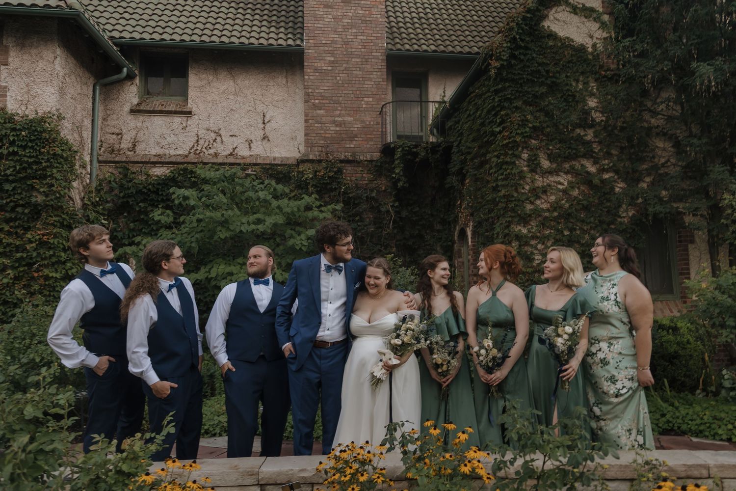Wedding party poses in front of ivy-covered brick venue, groomsmen in navy suits and bridesmaids in sage green dresses.