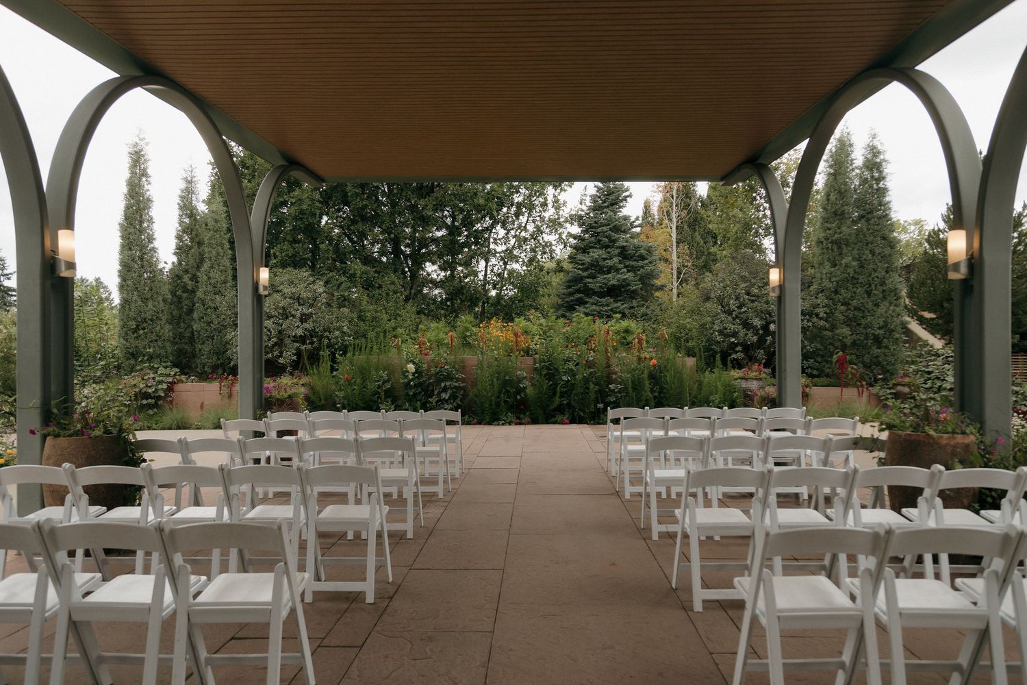 Outdoor wedding ceremony venue with white folding chairs under an elegant pergola surrounded by lush greenery and warm lighting.