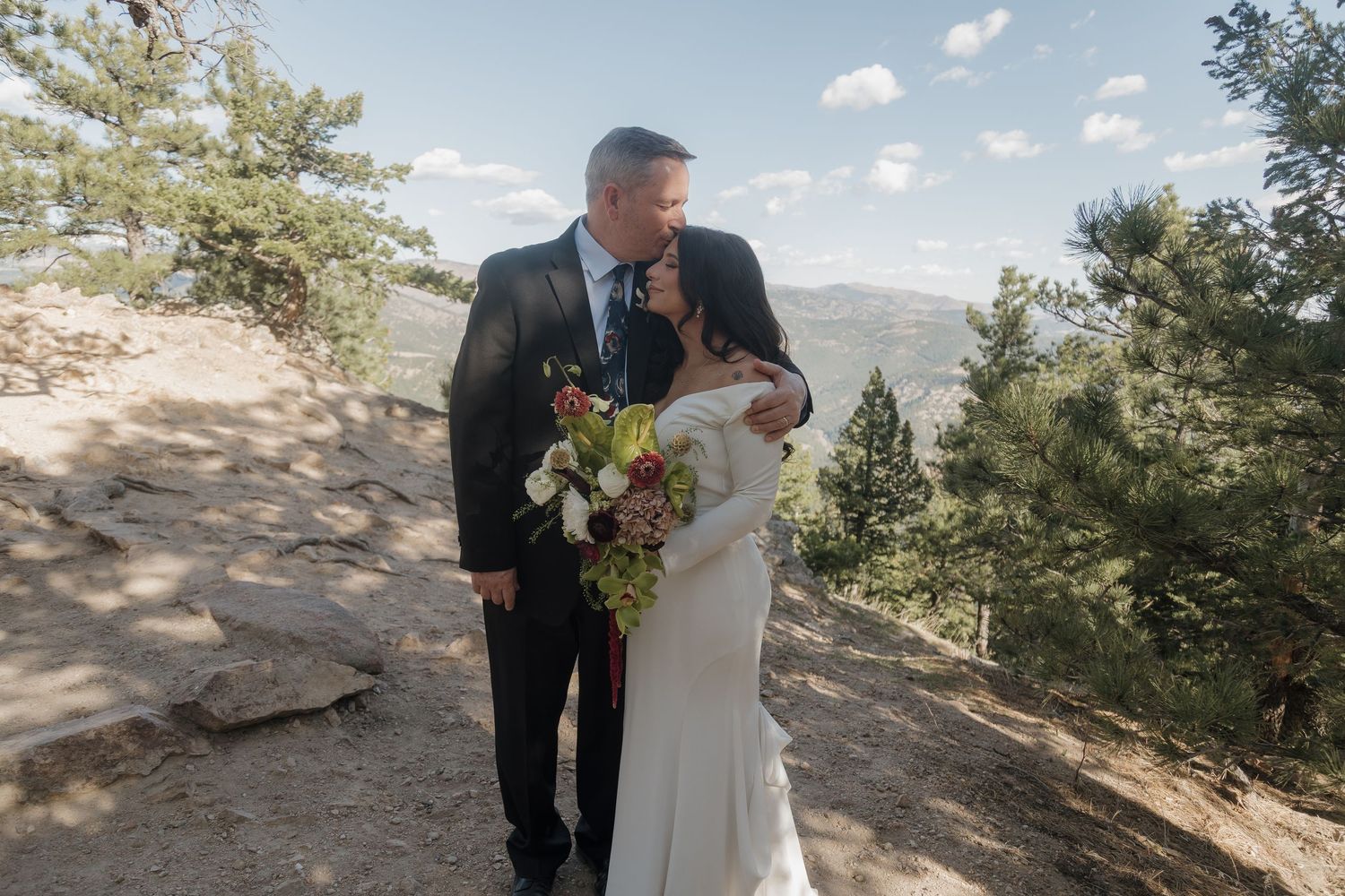Father of the bride embraces his daughter holding a floral bouquet on a scenic mountain trail.