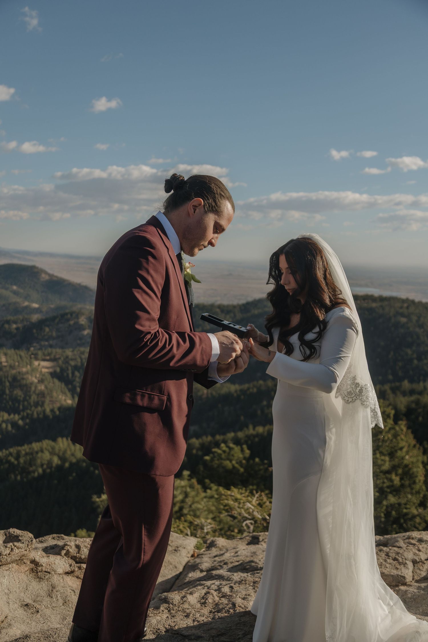 Groom in burgundy suit placing ring on bride's finger during outdoor mountain elopement ceremony.