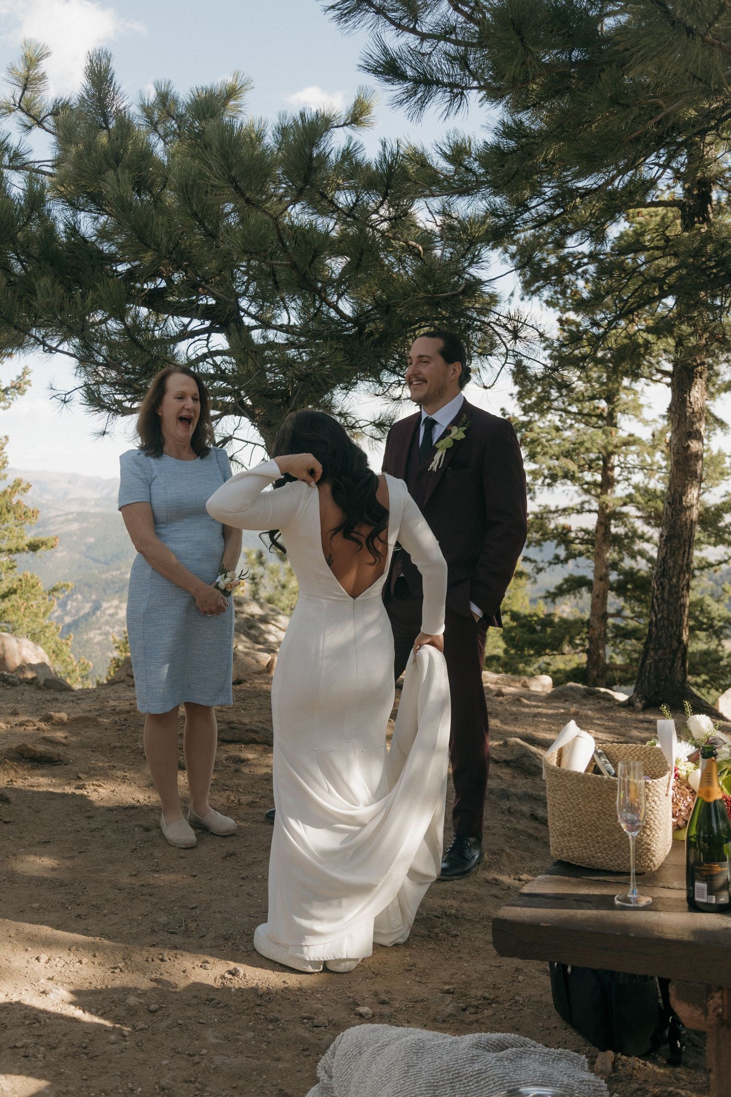 Bride laughing emotionally during outdoor mountain wedding ceremony with groom and officiant among pine trees.