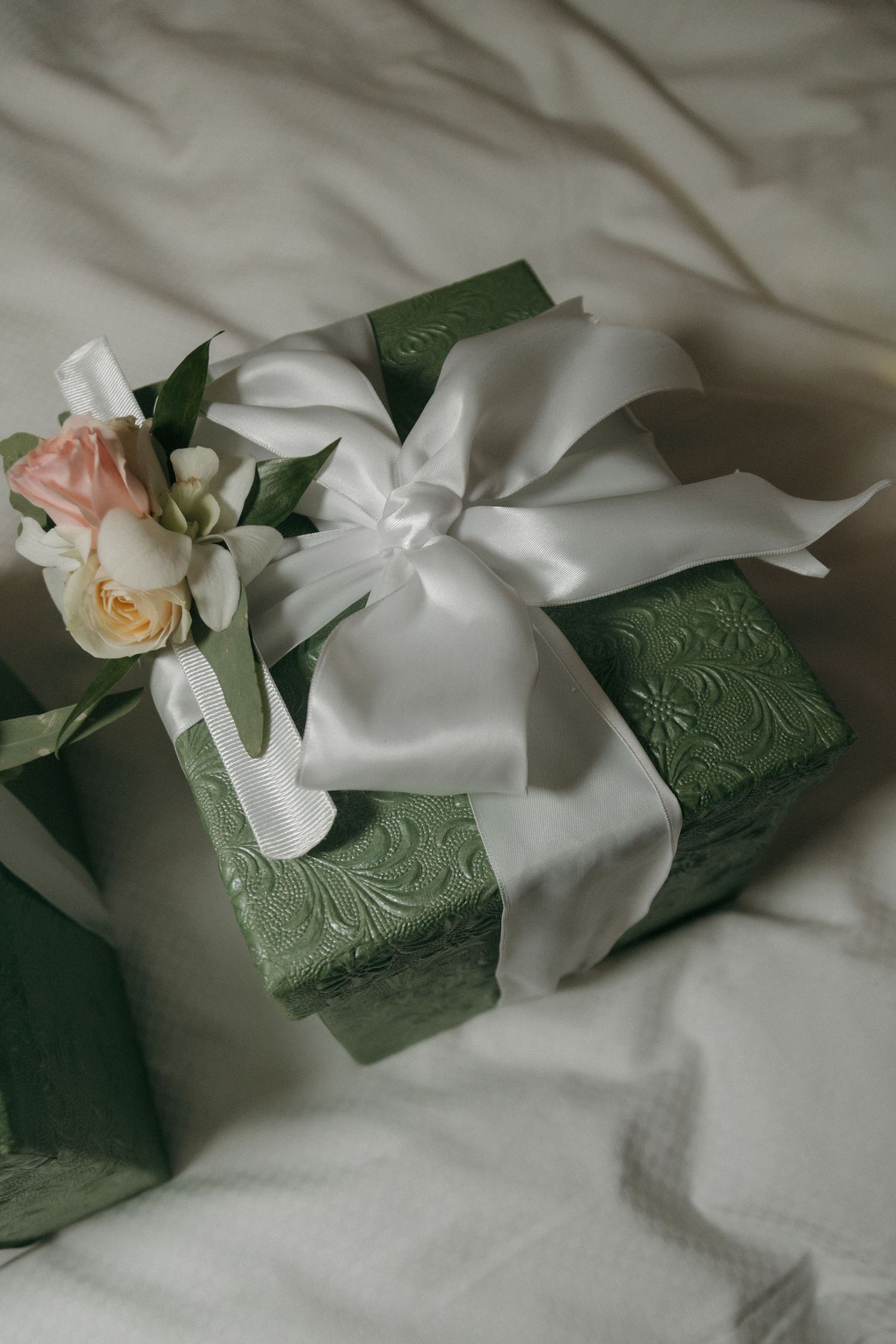 Elegant green embossed gift box with white satin bow and pink roses on white fabric.
