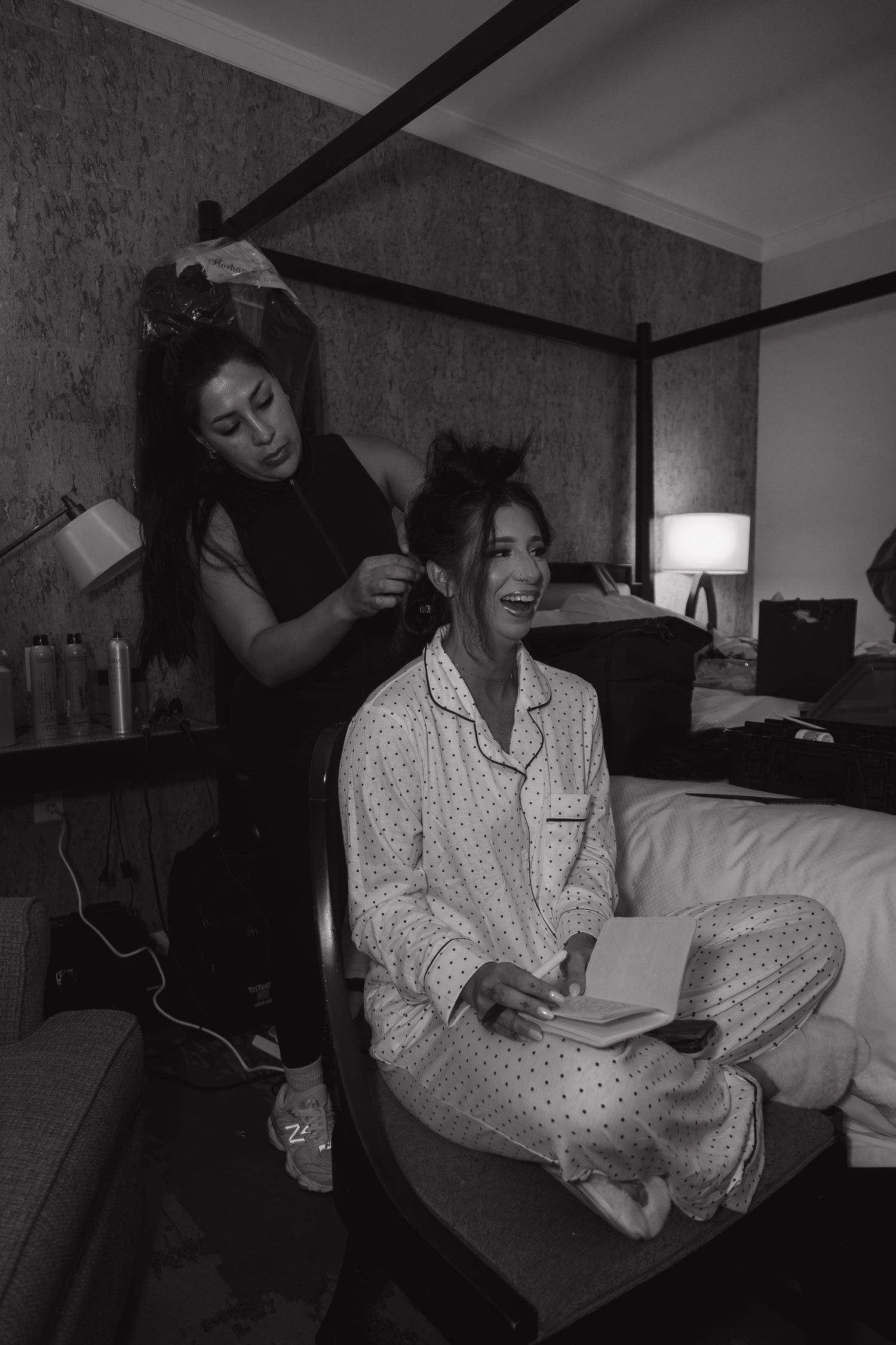 Black and white photo of bride laughing in polka dot pajamas while hairstylist works on her hair in hotel room.