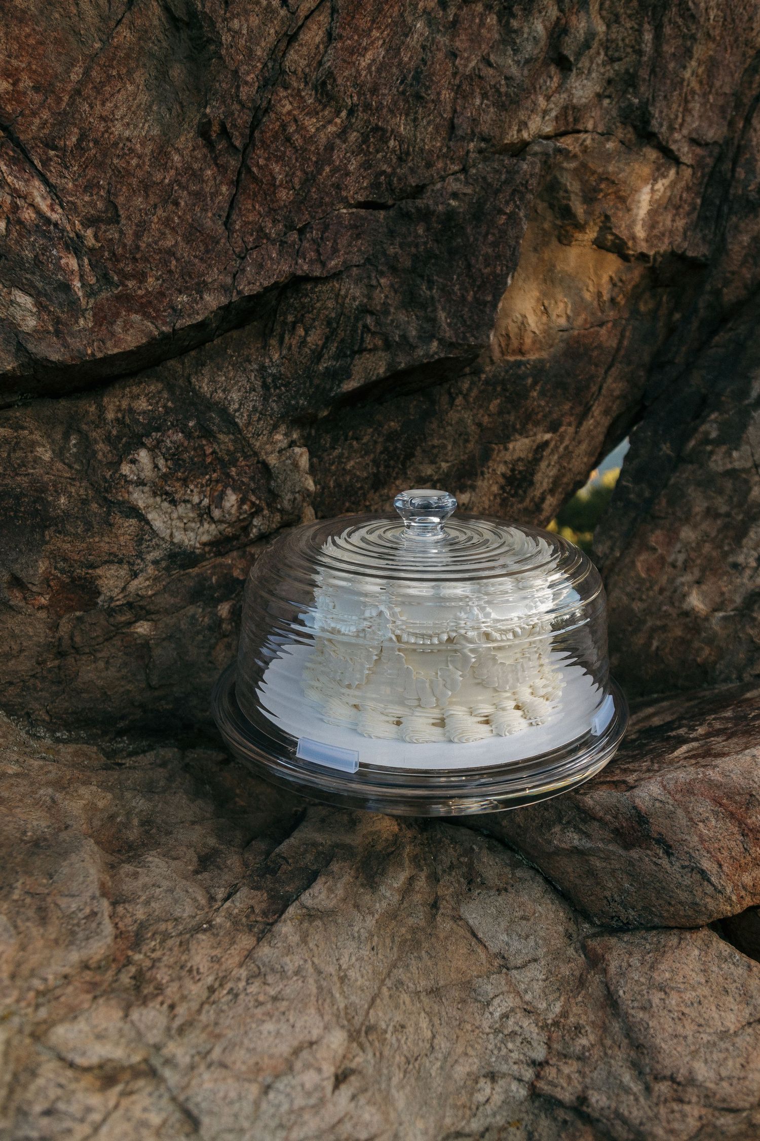 A glass cake dome displayed on rugged rocky terrain at an outdoor wedding venue.
