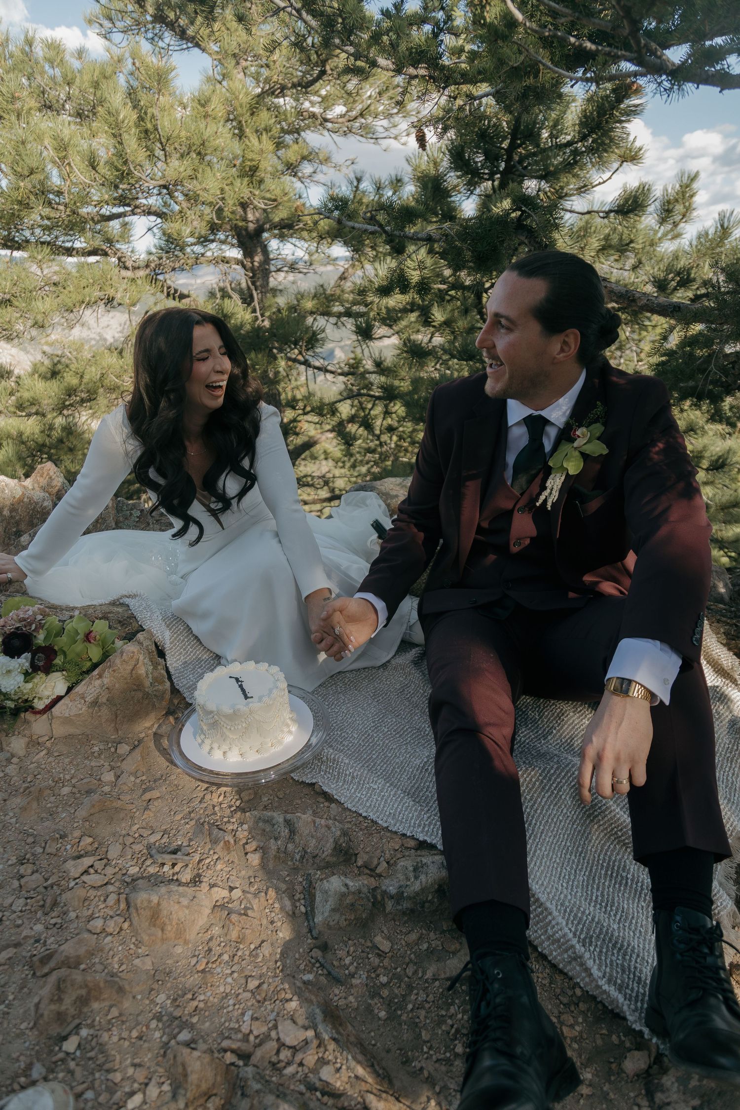 Bride and groom laugh joyfully while seated on rocks with a small white wedding cake beside them.