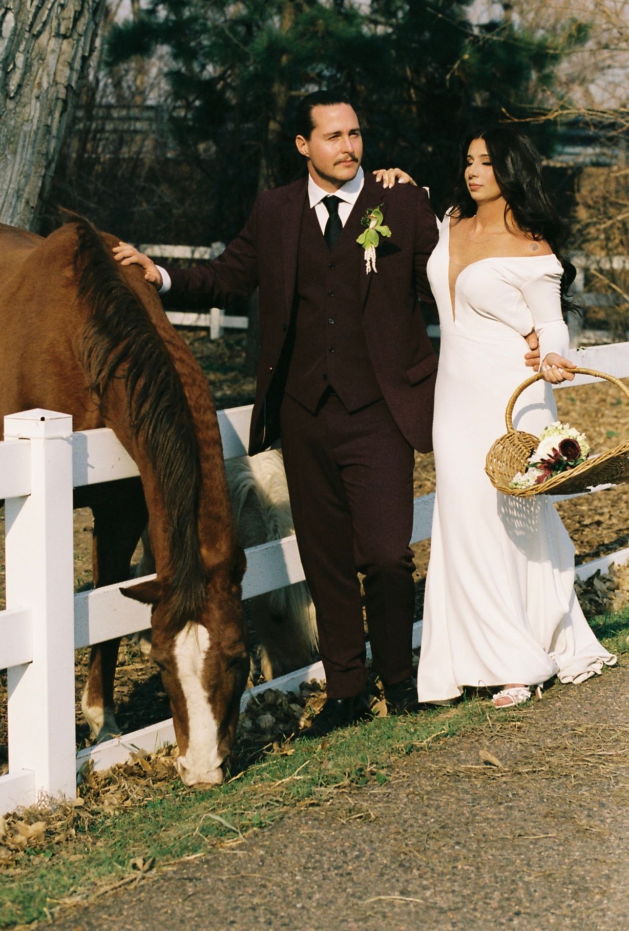 Bride in white off-shoulder gown and groom in dark suit petting horse by white fence at farm wedding.