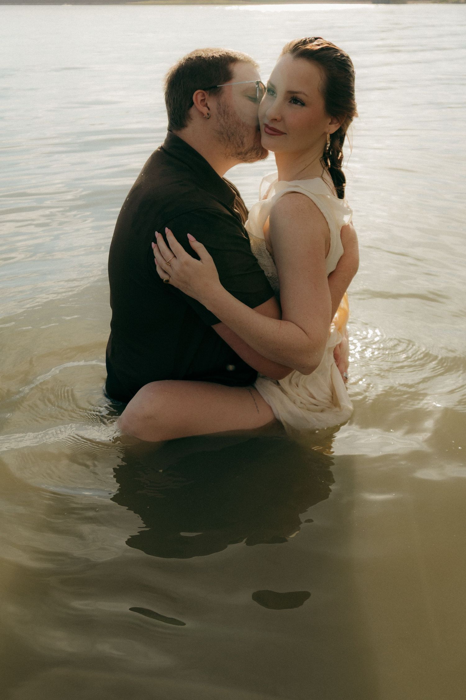 Couple embracing and kissing in lake water at sunset, bride in white dress and groom in black shirt.