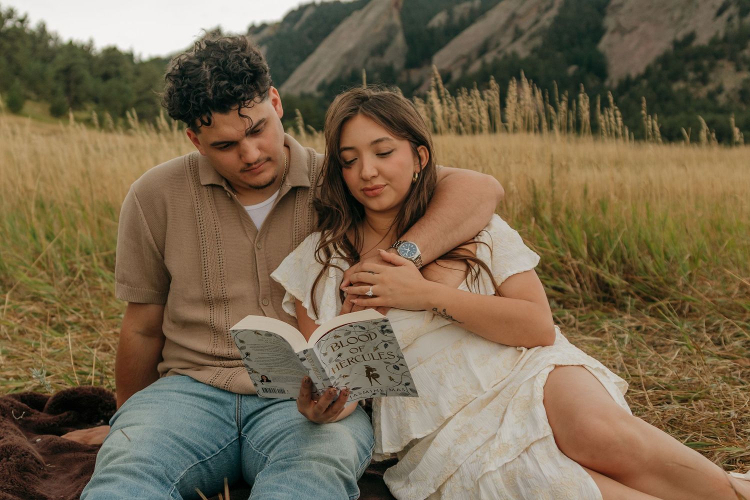 Couple sitting on blanket in field reading a book together, woman in white dress leaning on man.