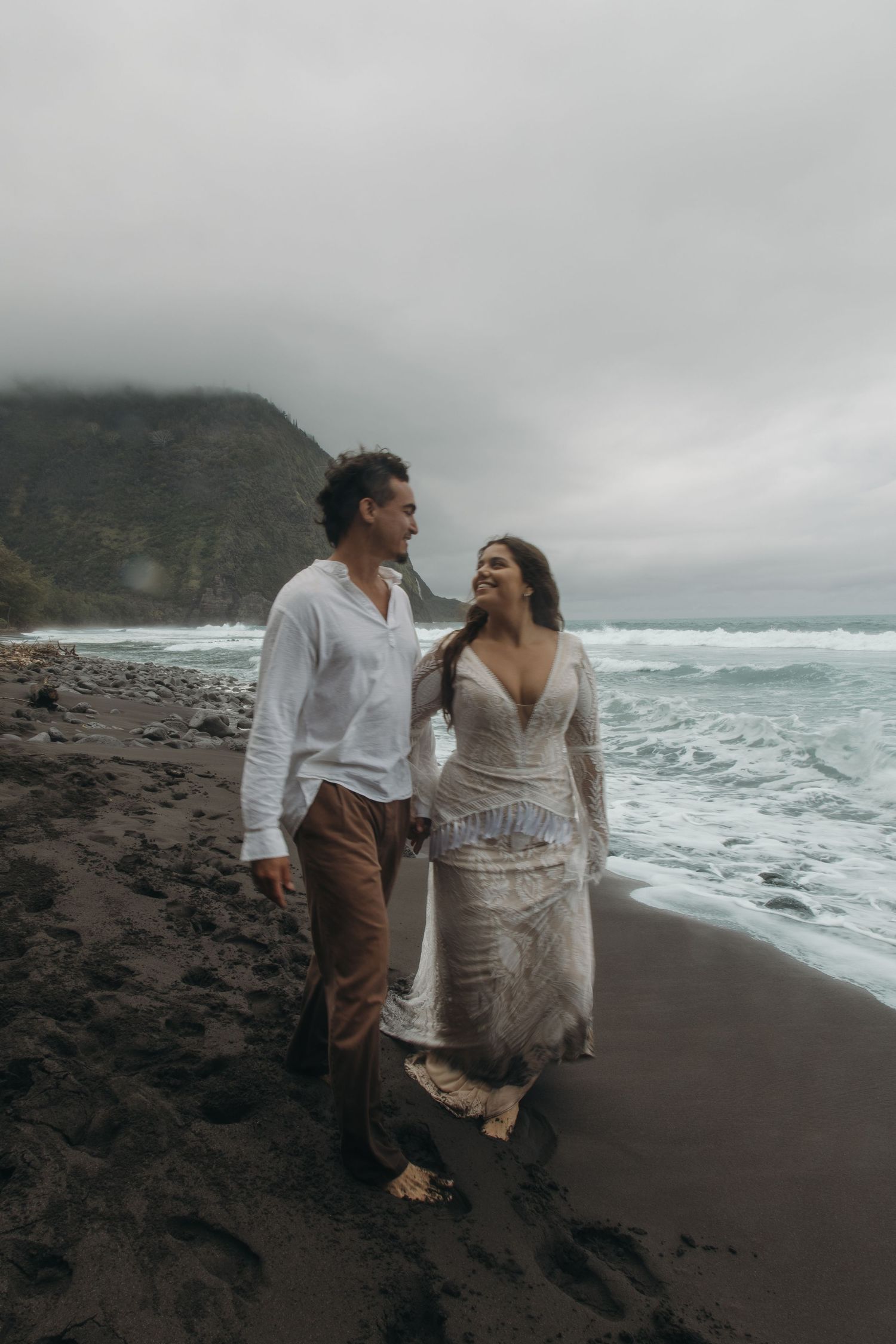 Smiling couple walk barefoot on black sand beach in white boho attire with dramatic cloudy skies.