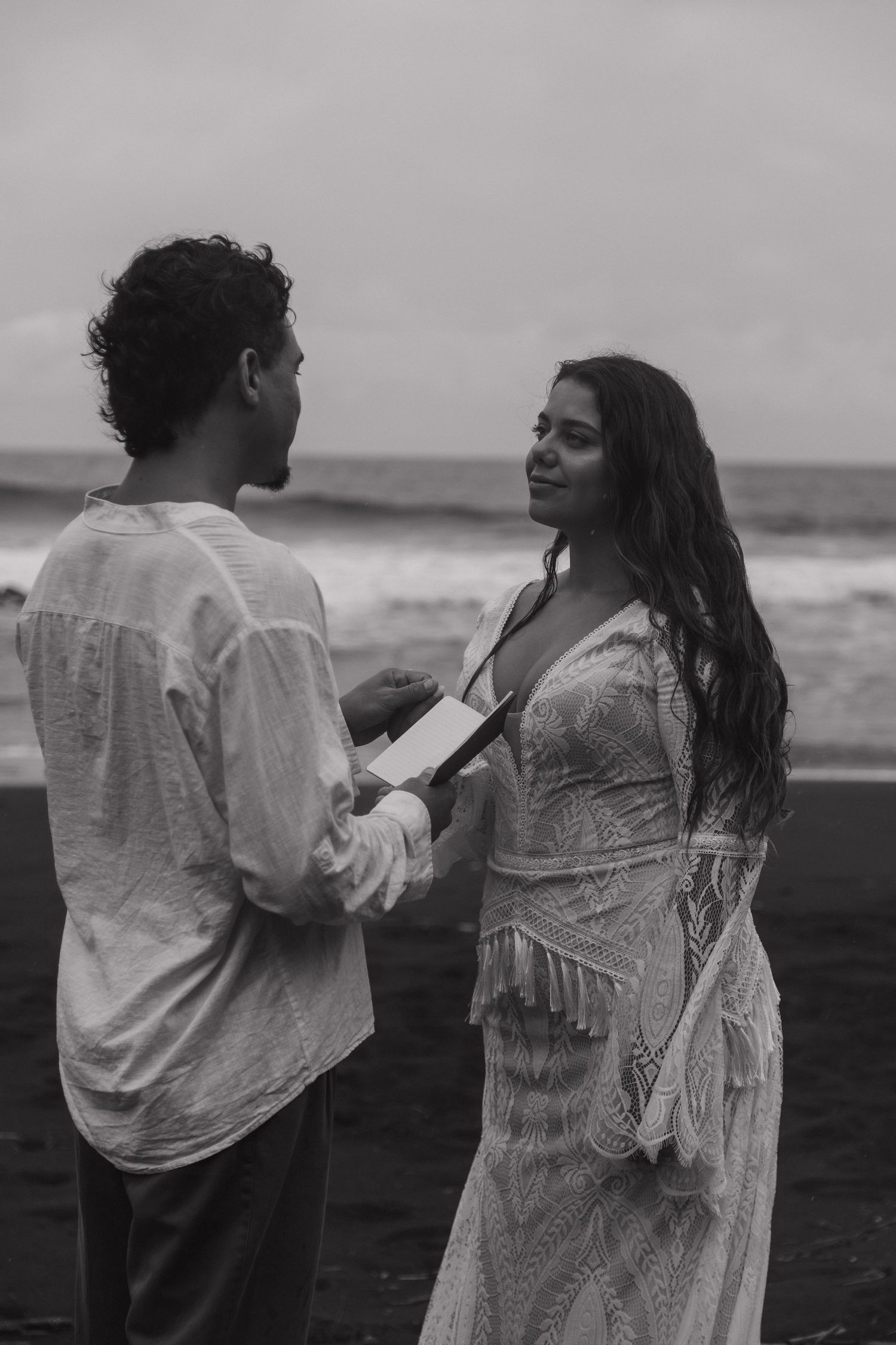 Black and white photo of couple exchanging vows on a black sand beach with ocean waves behind.