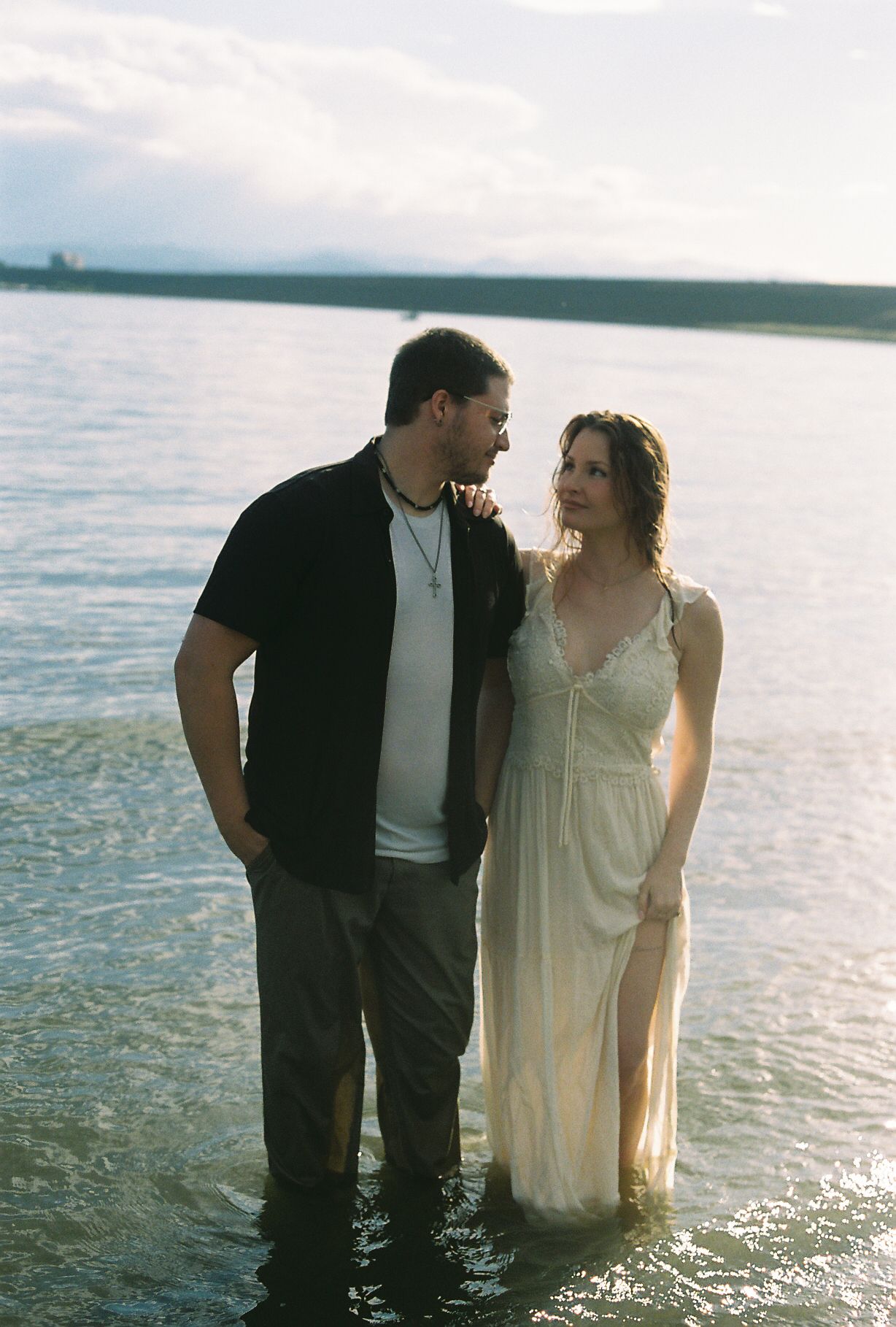 Couple standing together in shallow lake water, woman in flowing white lace dress at golden hour.