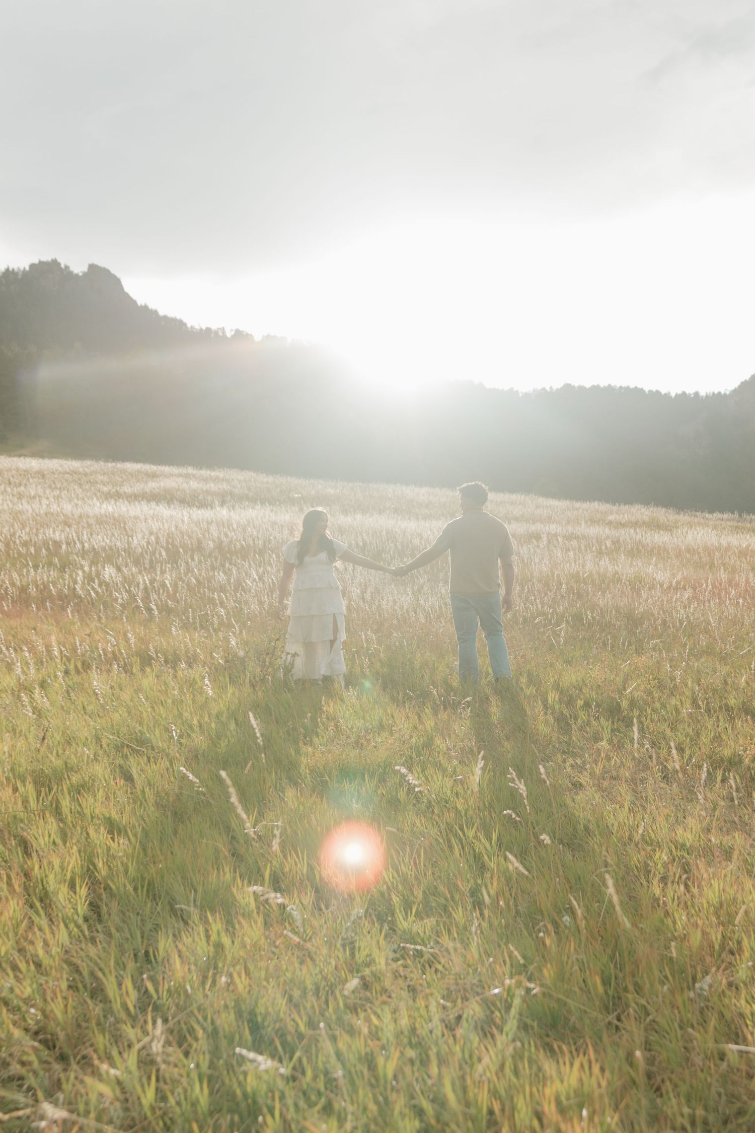 Couple holding hands in a sunlit golden field with misty mountains in the background.