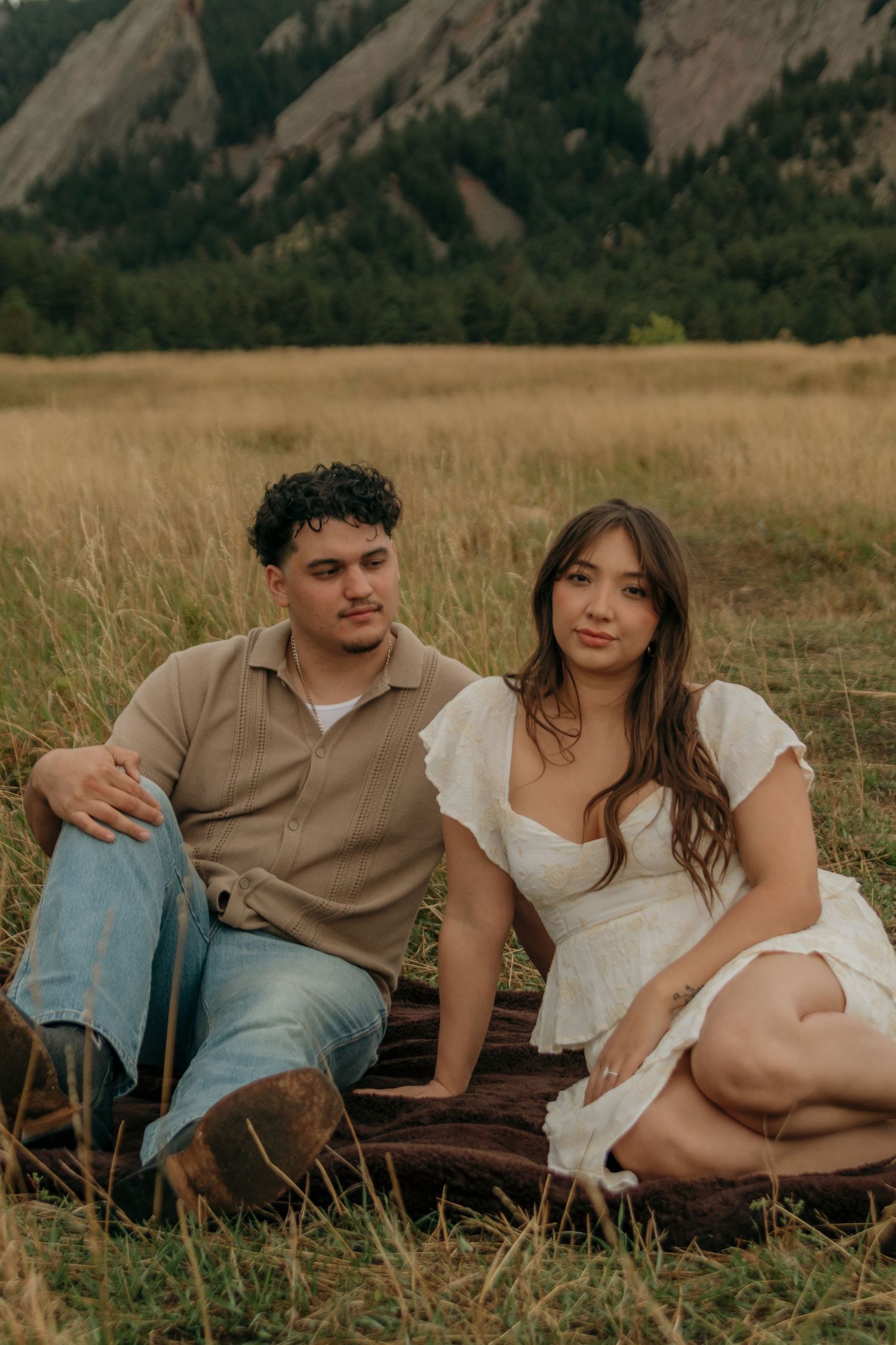 Couple sitting together in a grassy field with dramatic rocky mountain flatirons behind them.