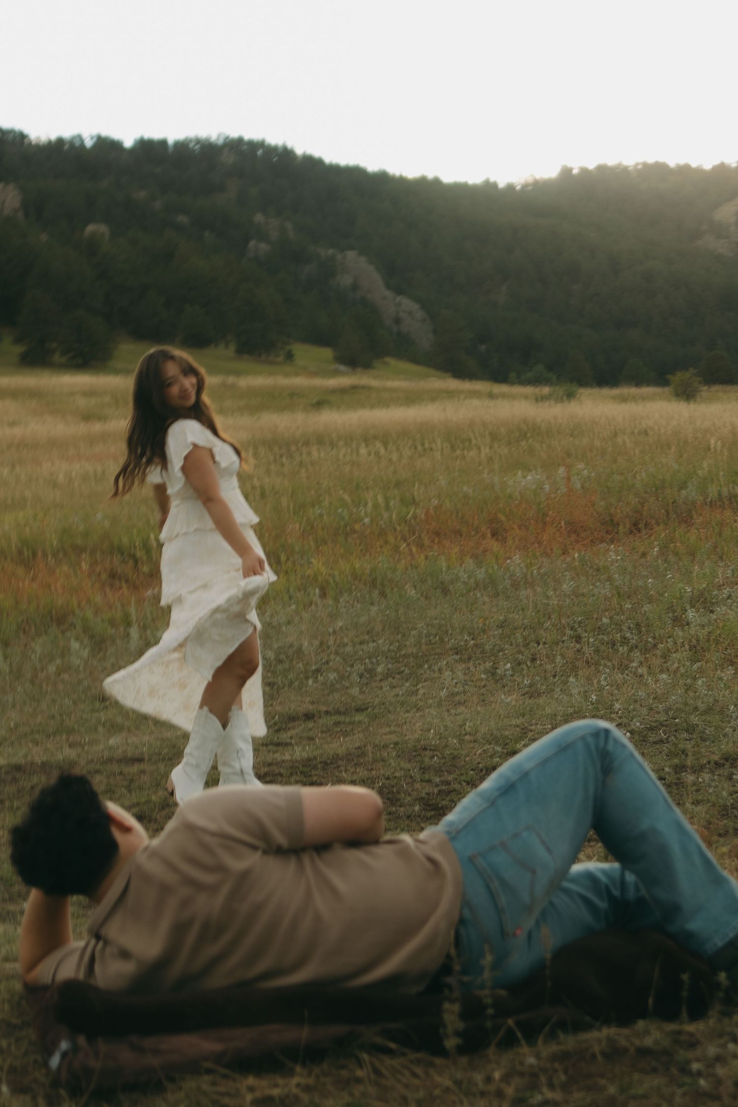 Woman in white dress smiling at camera while man relaxes in a sunlit mountain meadow.
