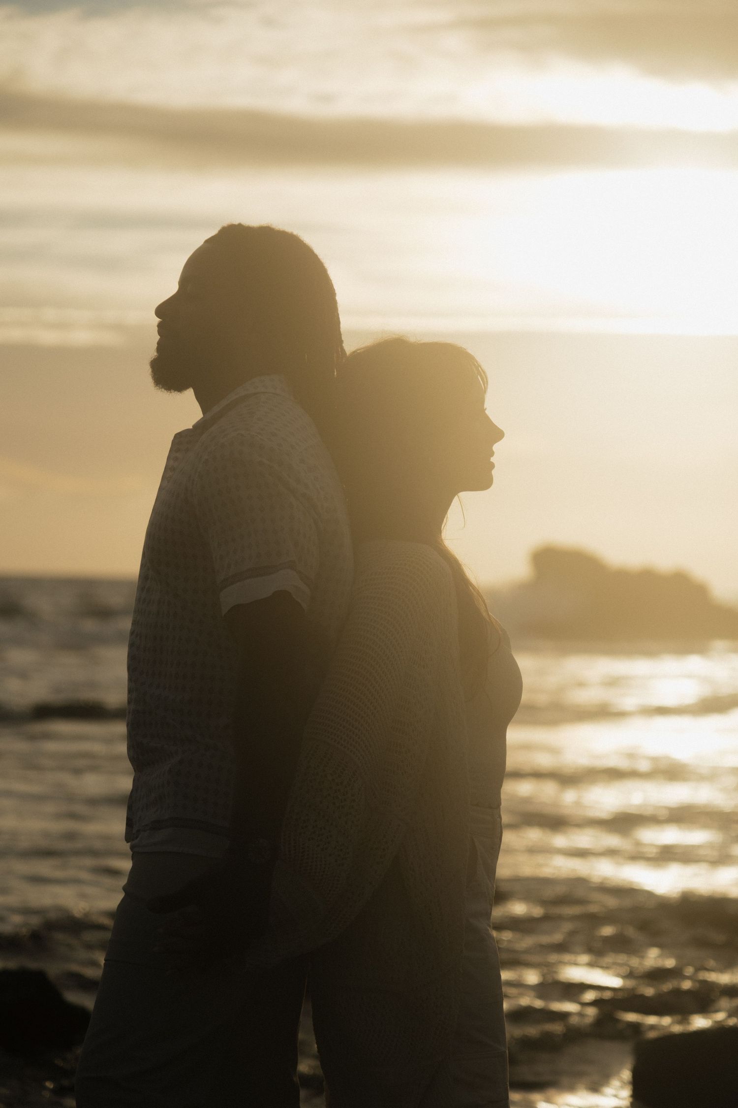 Silhouette of a couple standing back-to-back on rocky shore at golden sunset with ocean waves in background.