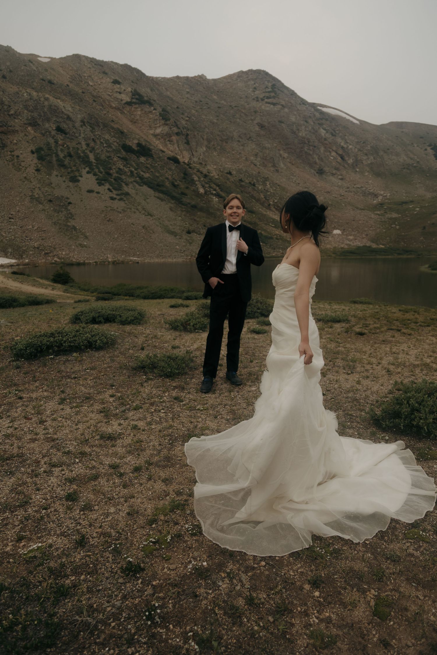 Bride in flowing white gown and groom in black tuxedo share a moment near a mountain lake during their outdoor elopement.