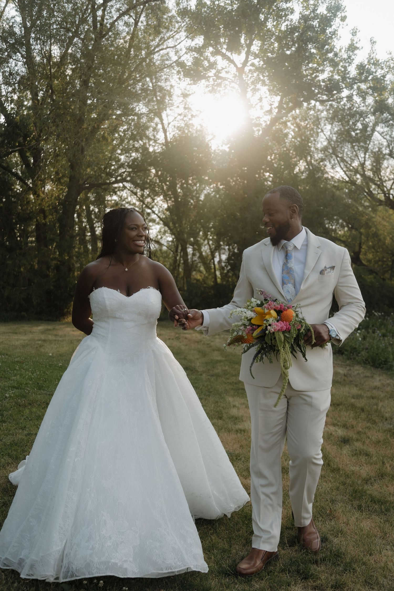 Newlyweds walk hand-in-hand at golden hour, groom in cream suit holding bright bouquet, bride in strapless white gown.