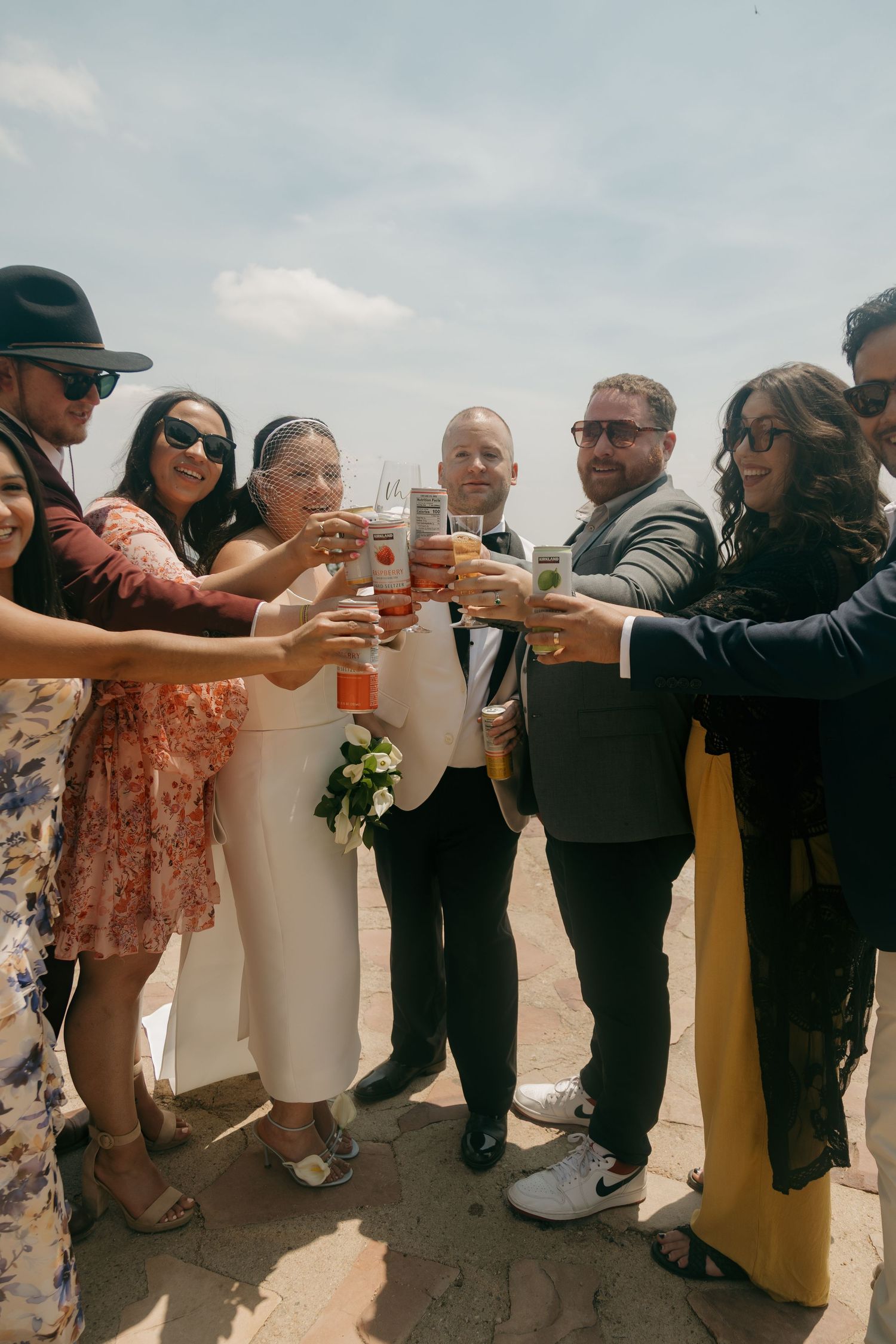 Wedding party raises glasses in celebratory toast outdoors under blue sky on wedding day.