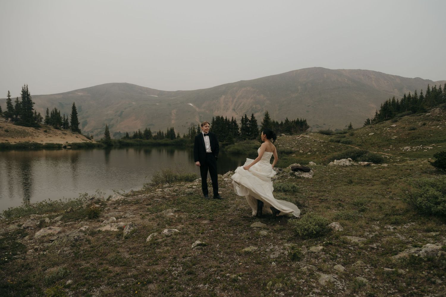 Bride twirls her white gown near a calm mountain lake while groom watches in a black tuxedo on an overcast day.