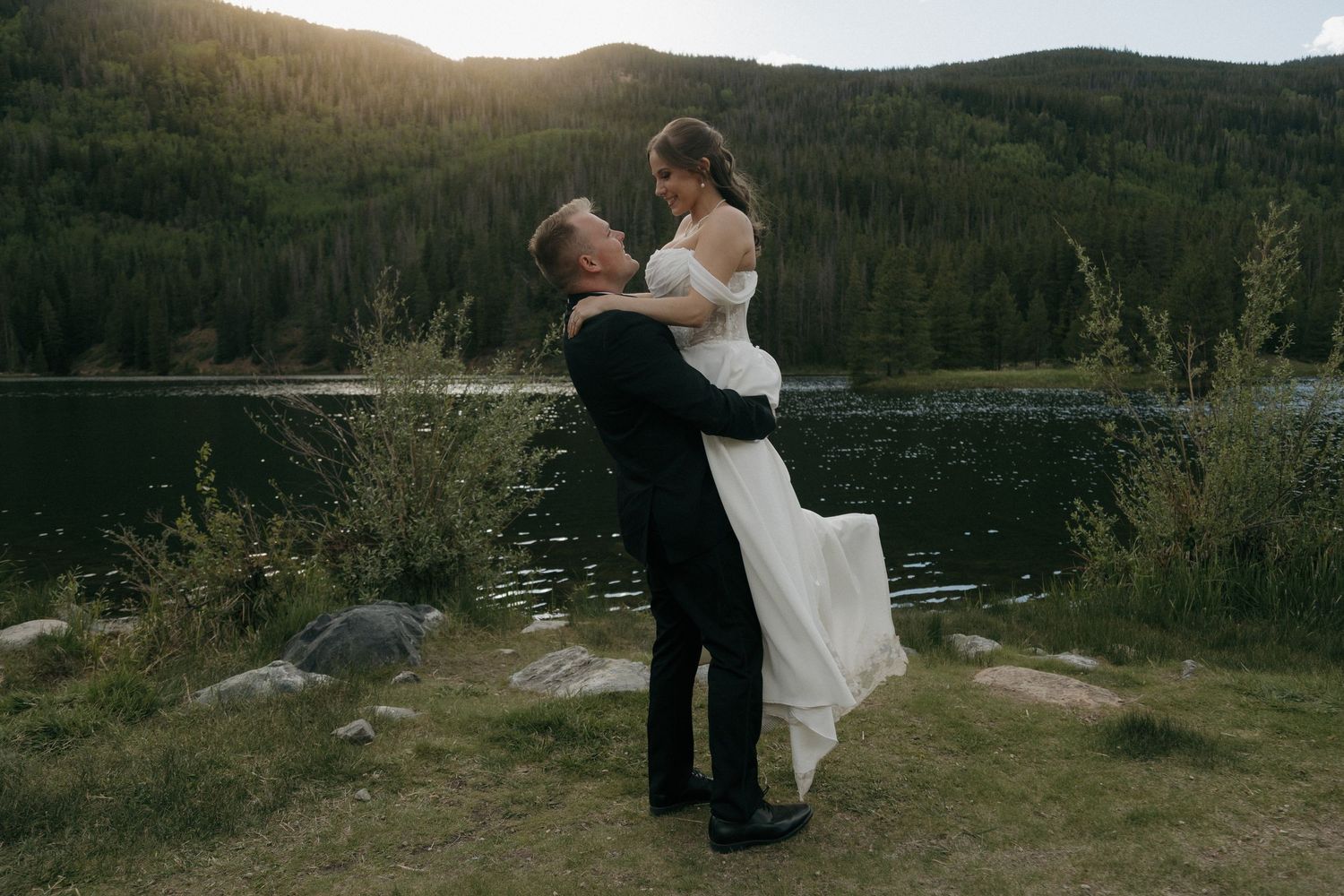 Groom lifting bride in white dress by a mountain lake at sunset during outdoor wedding celebration.