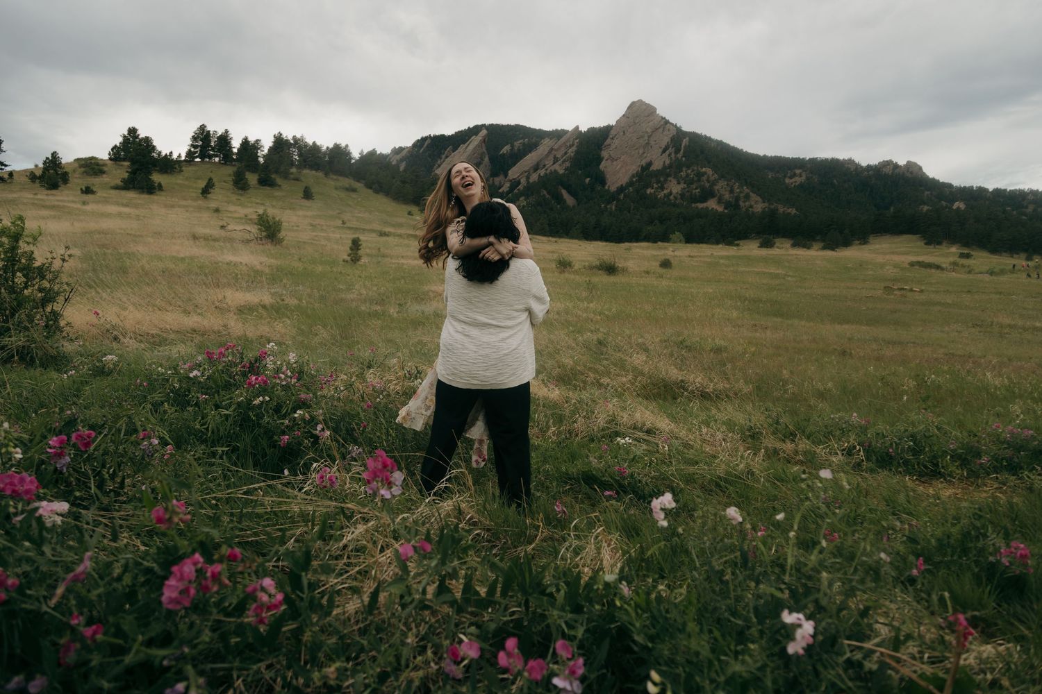 Couple embracing among pink wildflowers in a green meadow with the Flatirons mountains backdrop.