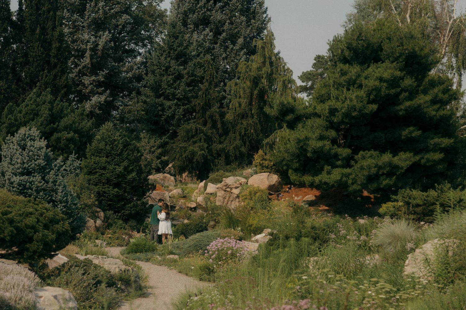 Woman in a white dress walking alone on a winding path through a lush rocky botanical garden.