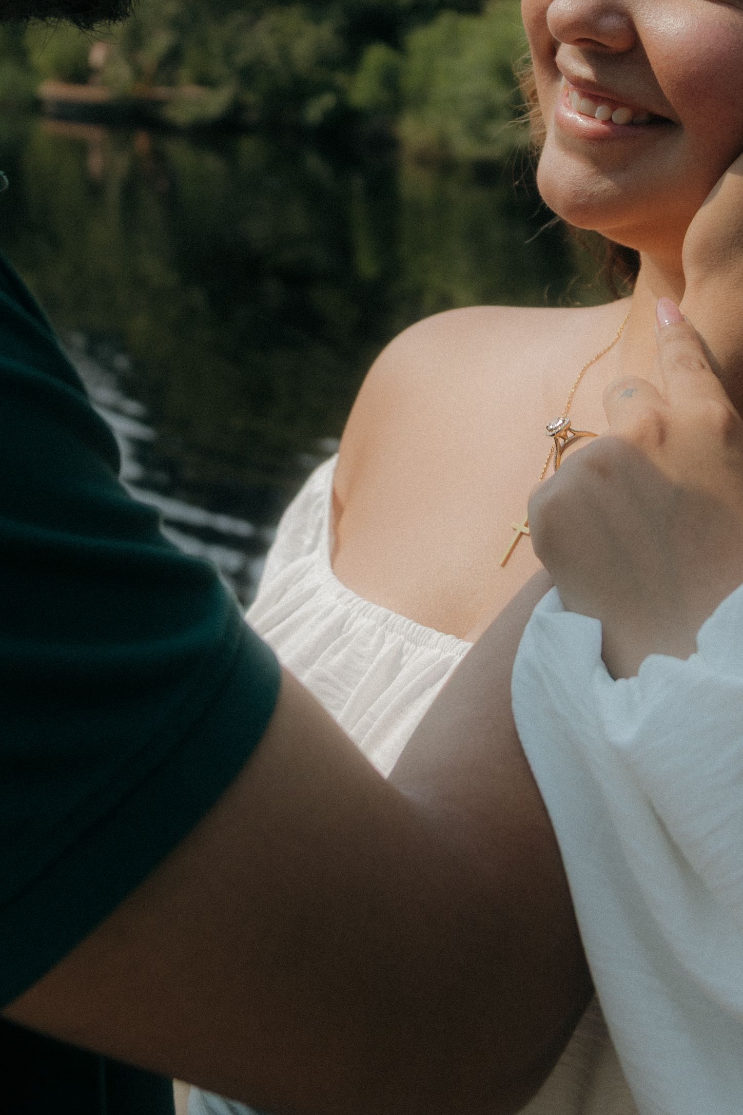 Smiling woman in white off-shoulder dress holding a gold necklace near a serene lake backdrop.