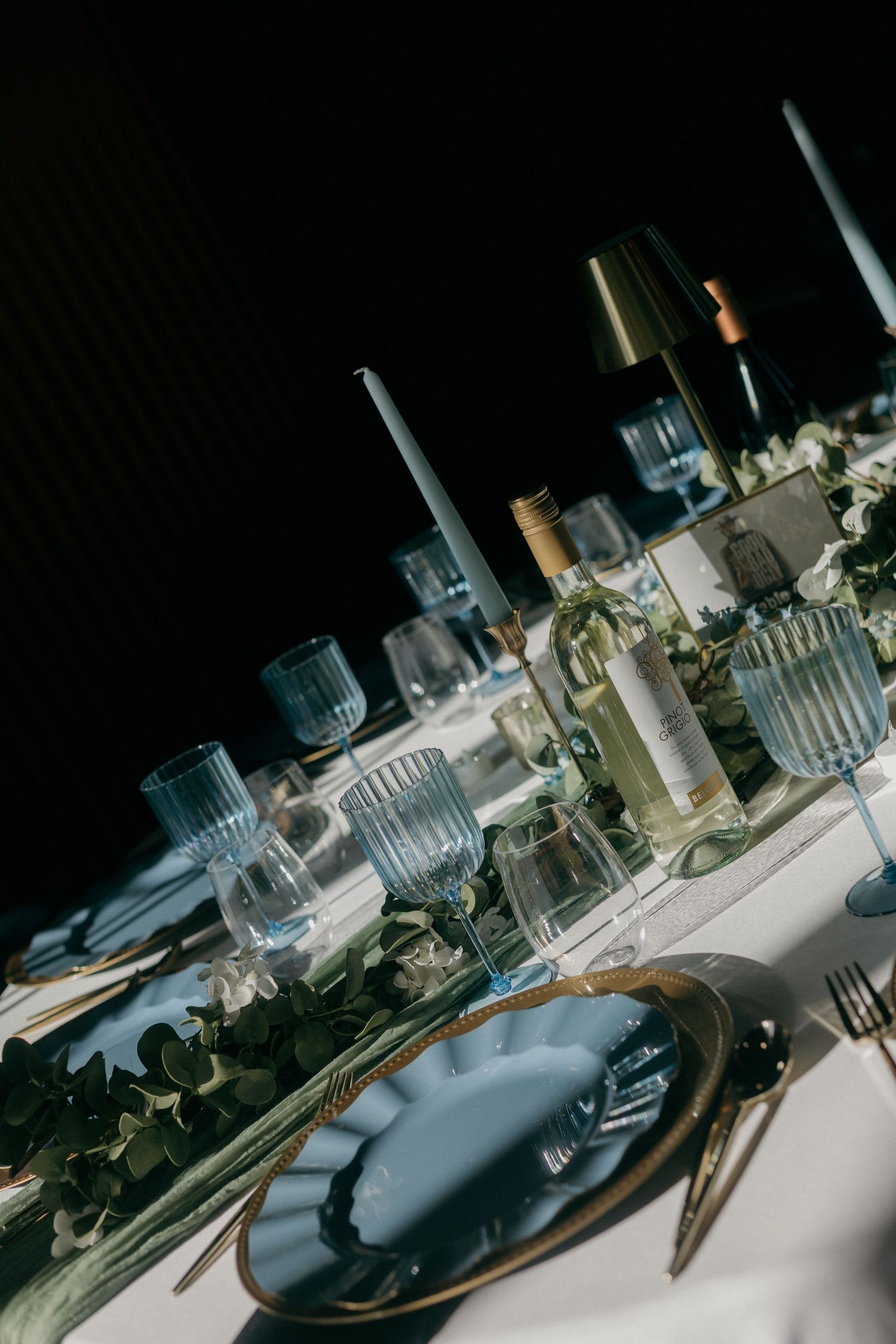 Elegant wedding table setting with blue glassware, taper candles, greenery garland, and gold-rimmed charger plates on white linen.