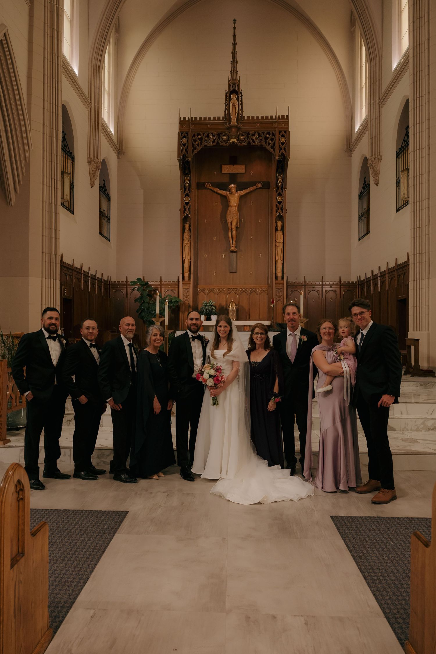 Wedding party poses with bride and groom before crucifix altar in Catholic church after ceremony.