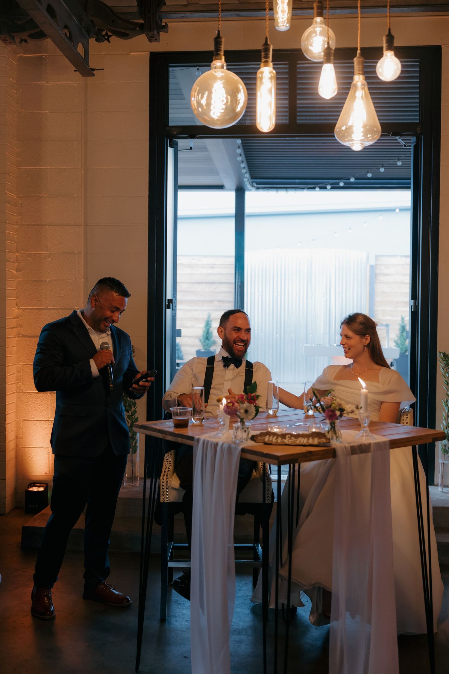 Bride and groom laughing at sweetheart table with Edison bulb lighting during intimate wedding reception.