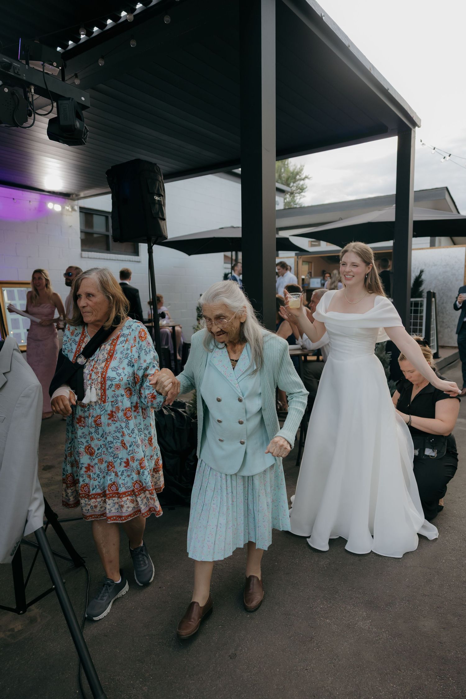 Bride in white gown dancing outdoors with elderly guests at sunny wedding reception.