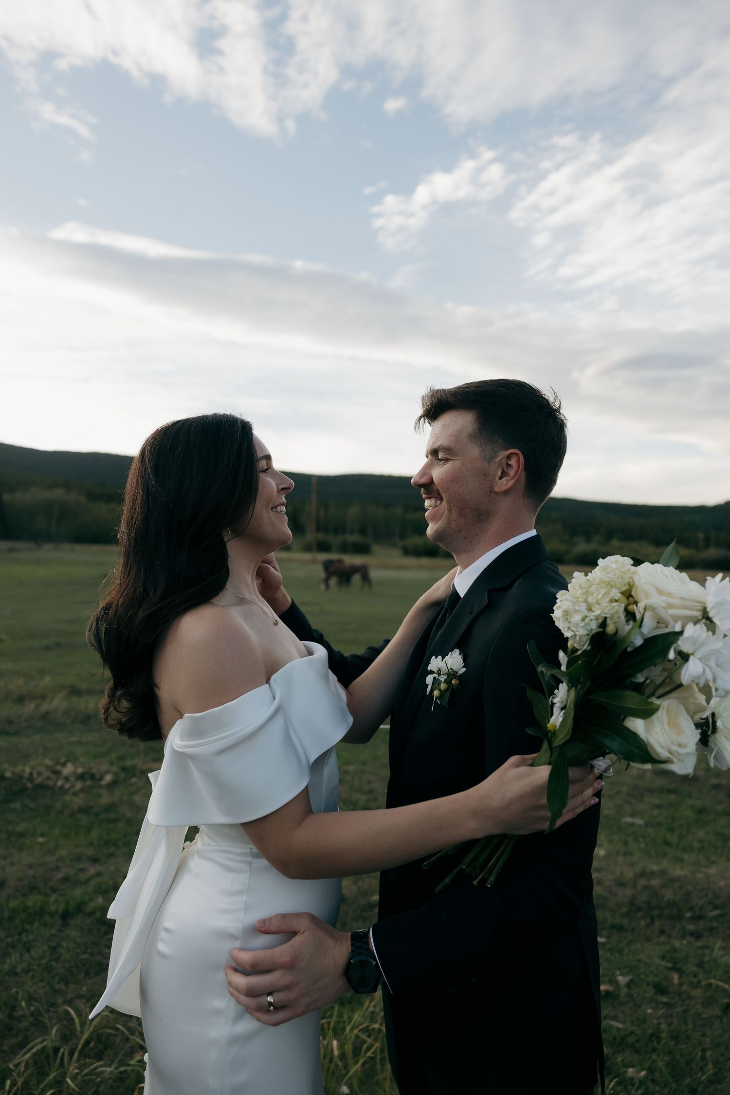 Bride and groom smiling at each other in field with horses grazing in background at sunset.