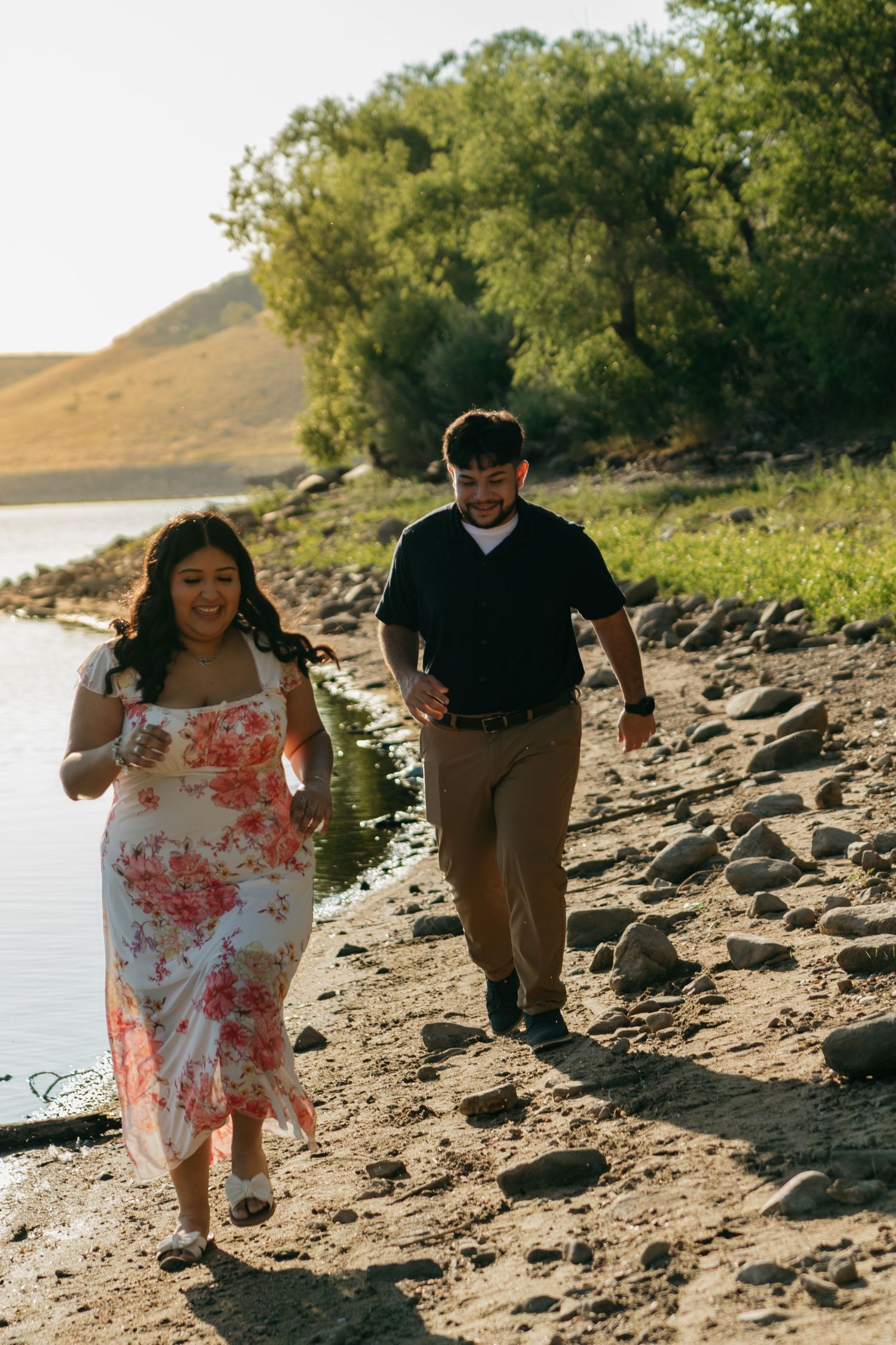 A couple runs playfully along a lakeside trail with trees in the background.