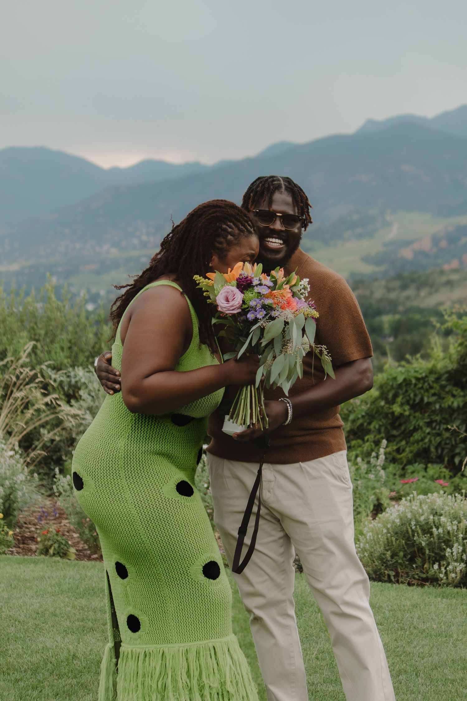 A couple shares a joyful moment with flowers against a mountain backdrop wearing green polka dot dress and brown shirt.