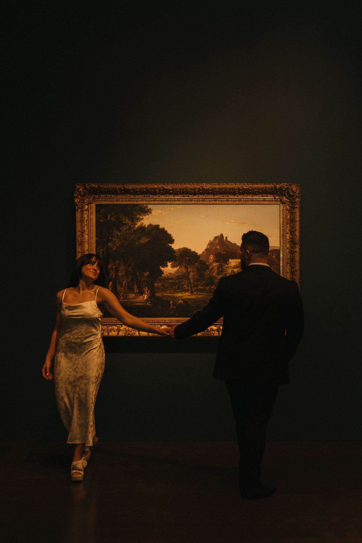 Figures stand in shadow viewing artwork displayed in golden frames on a dim museum wall.