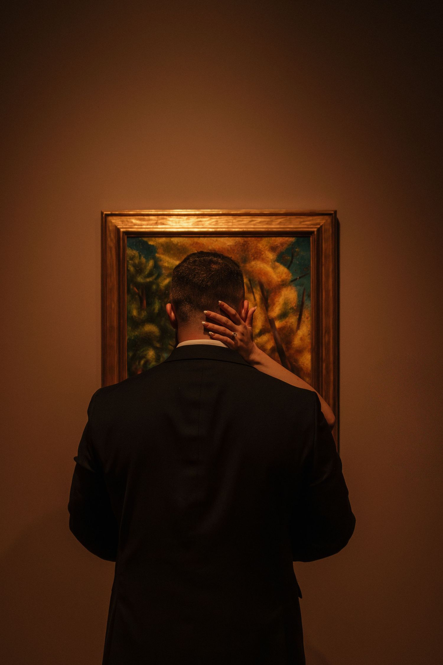 Silhouetted figures examine framed artwork on a dark brown gallery wall.