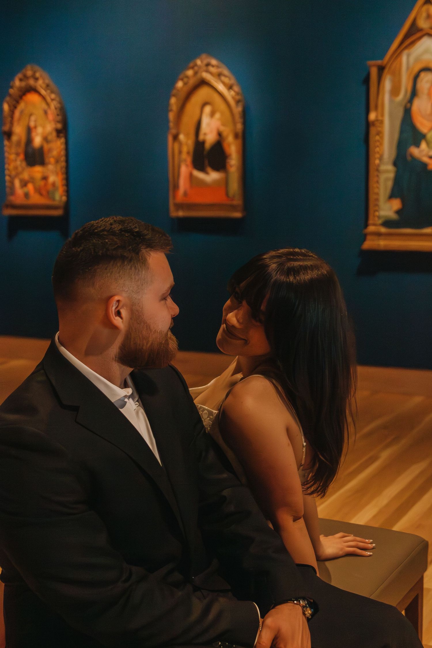 A couple shares an intimate moment on a bench in front of ornate religious art displays.