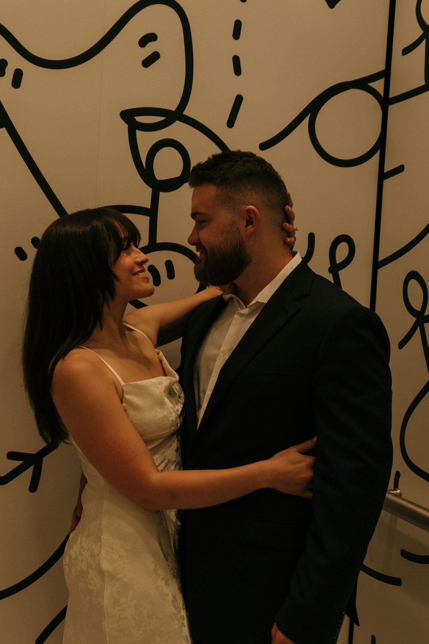 A couple in formal wedding attire shares an intimate moment against a decorative wall.