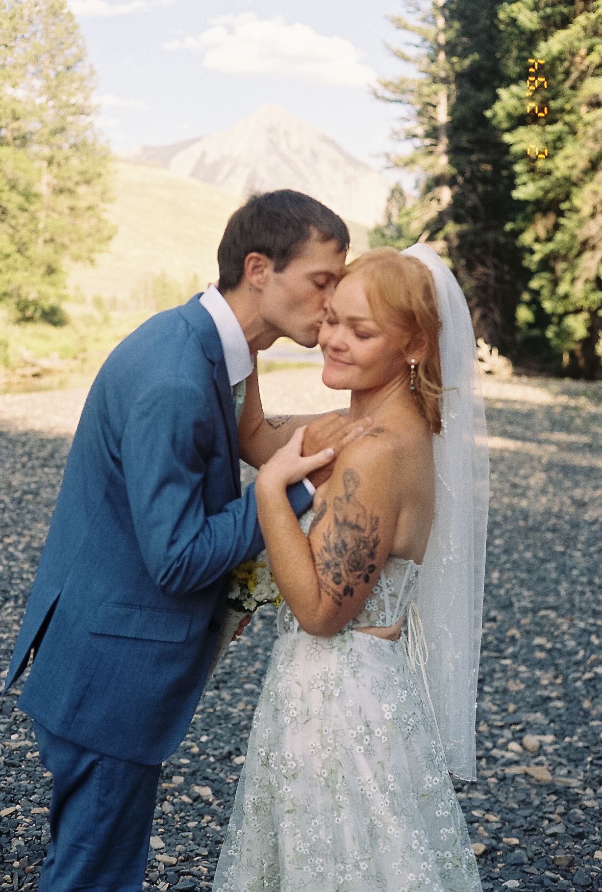 Groom in blue suit kissing bride on forehead, bride wearing lace gown with floral back tattoo near mountains.