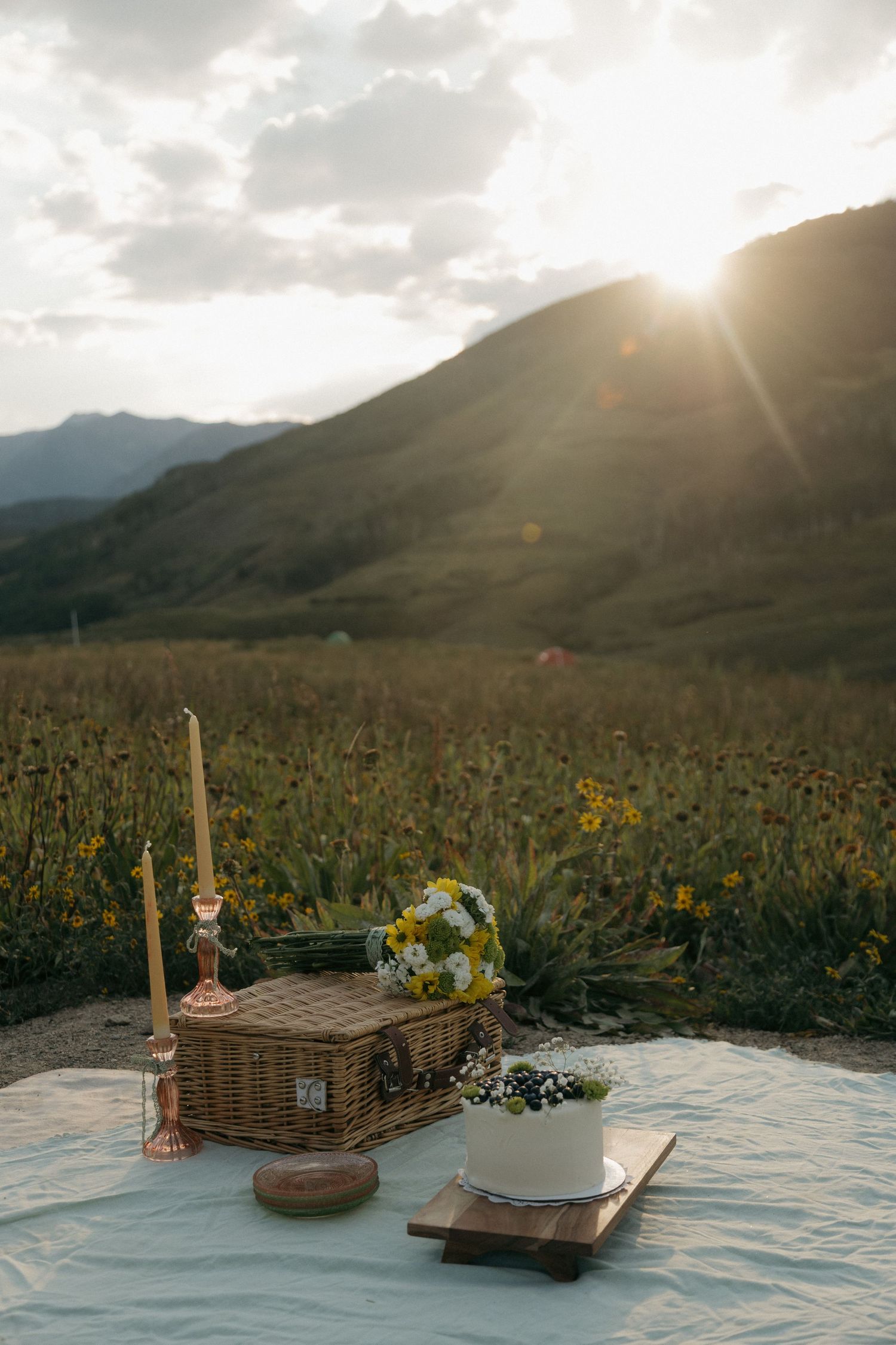 Rustic picnic setup with wicker basket, cake, and candle in a mountain wildflower meadow at golden sunset.