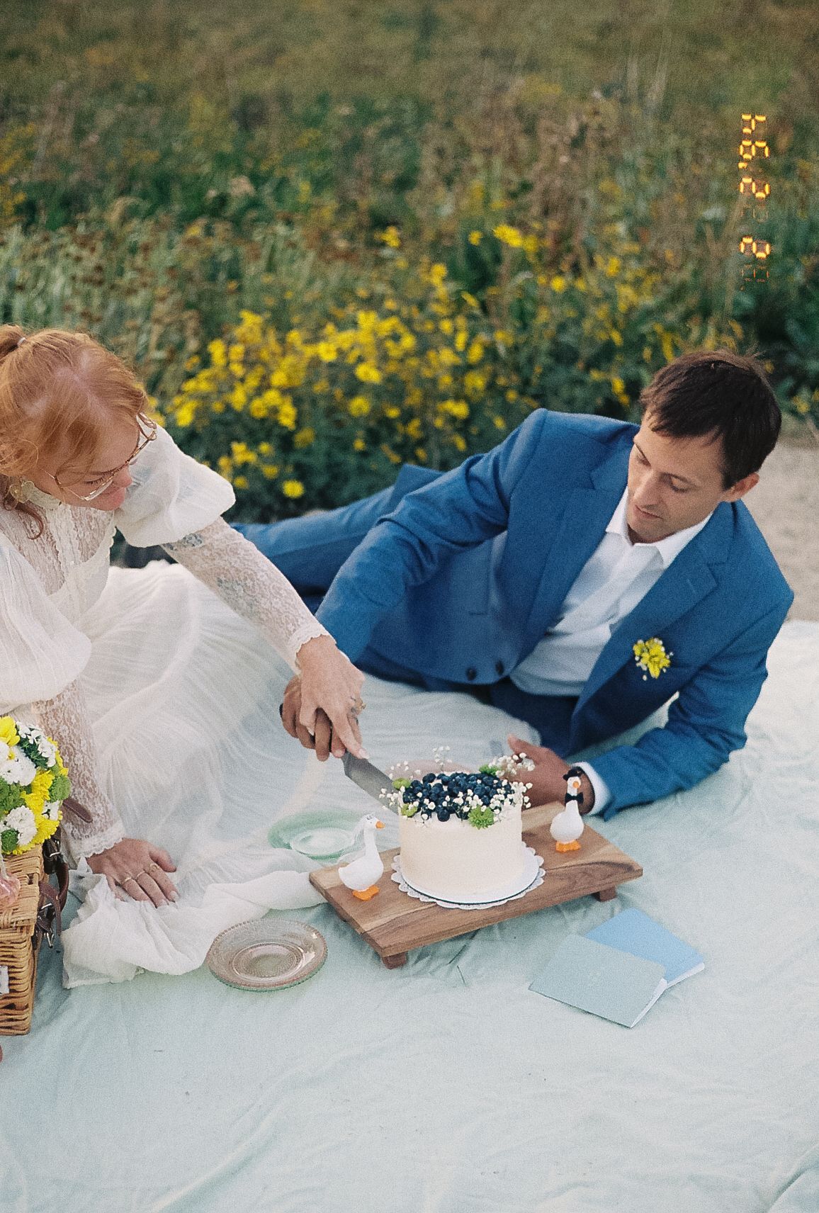 Bride and groom cutting white wedding cake on picnic blanket outdoors, surrounded by yellow wildflowers.