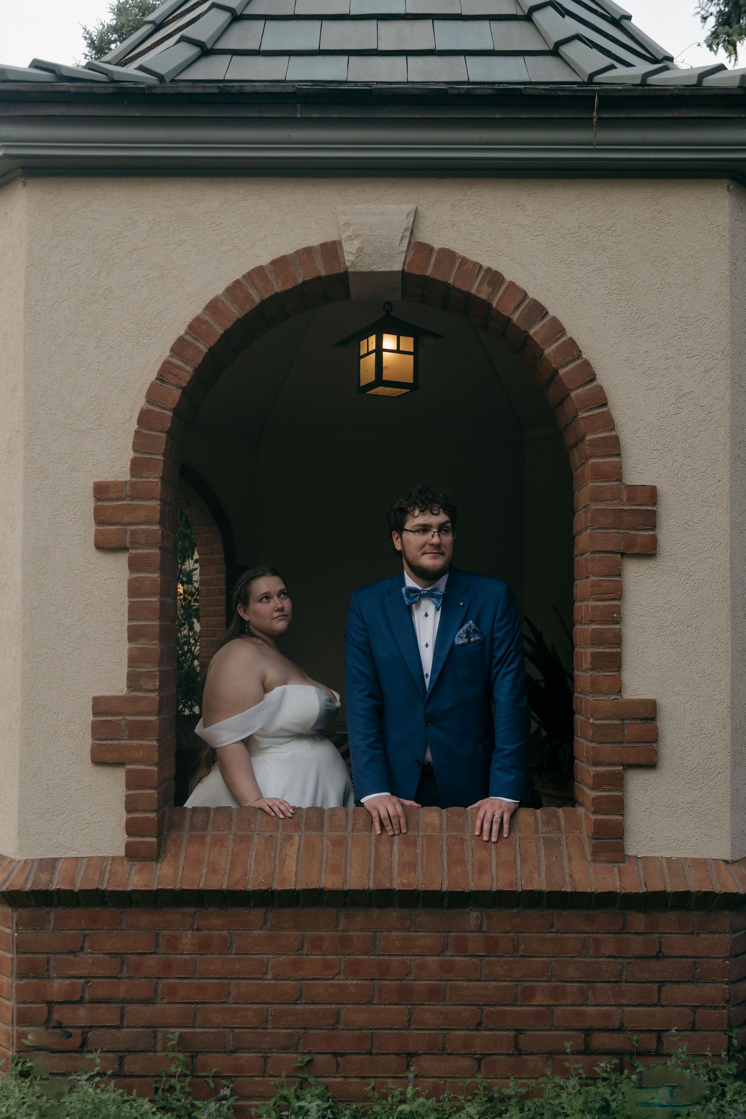 Bride and groom pose in arched brick gazebo window with lantern overhead at outdoor wedding.