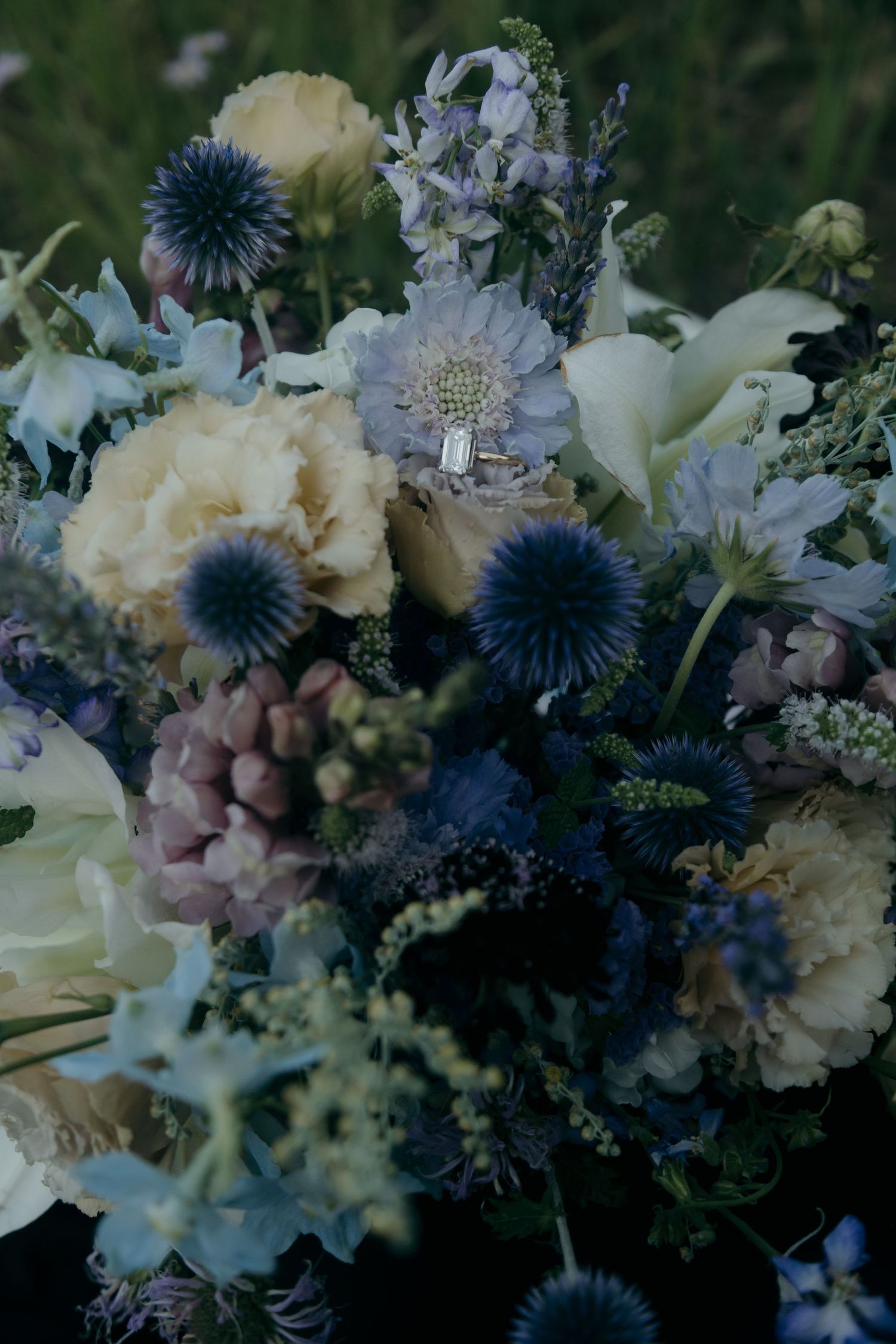 Elegant wedding bouquet featuring blue thistle, lavender, cream peonies, and wildflowers with diamond engagement ring nestled inside.