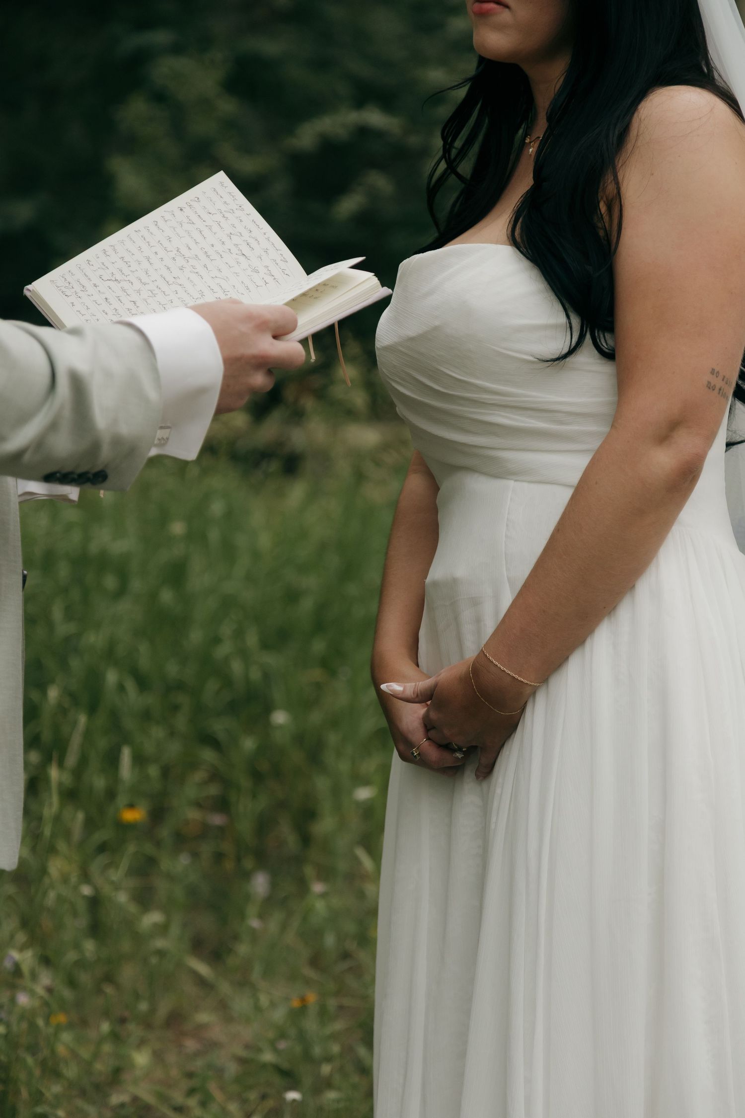 Bride in strapless white gown holds hands while groom reads wedding vows from a book during outdoor ceremony.