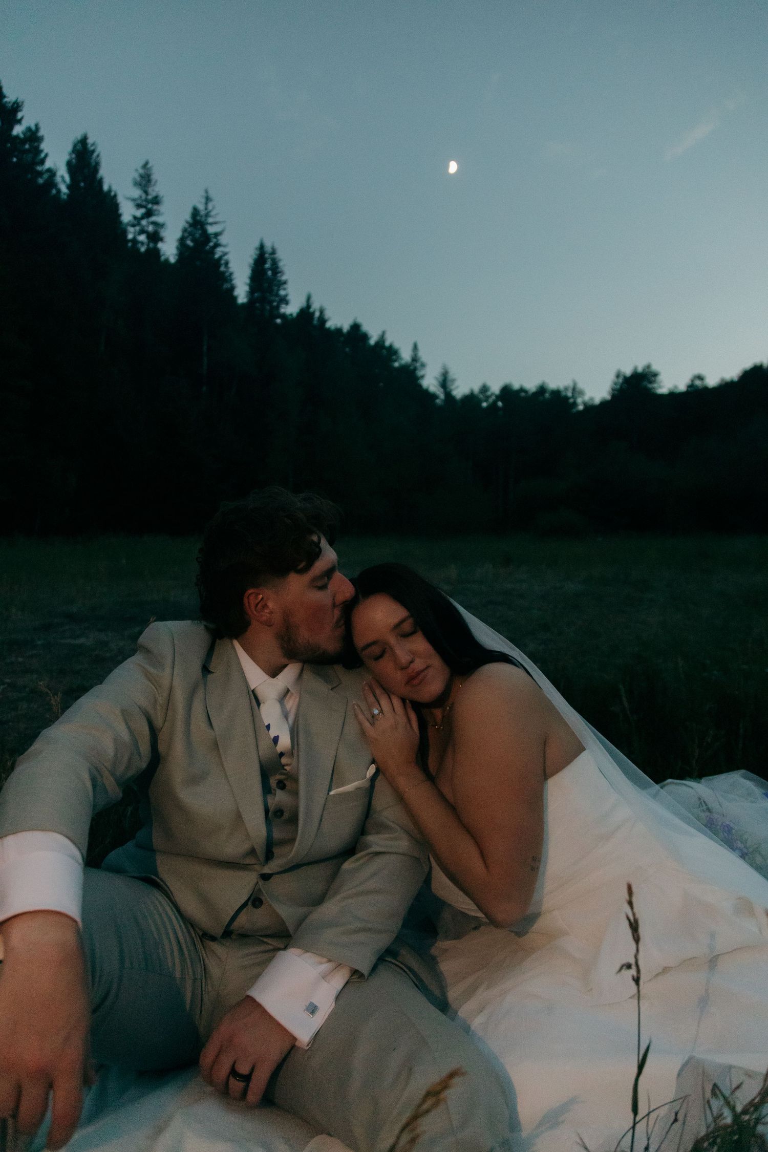 Romantic bride and groom sitting together under a moonlit sky in a serene forest meadow.