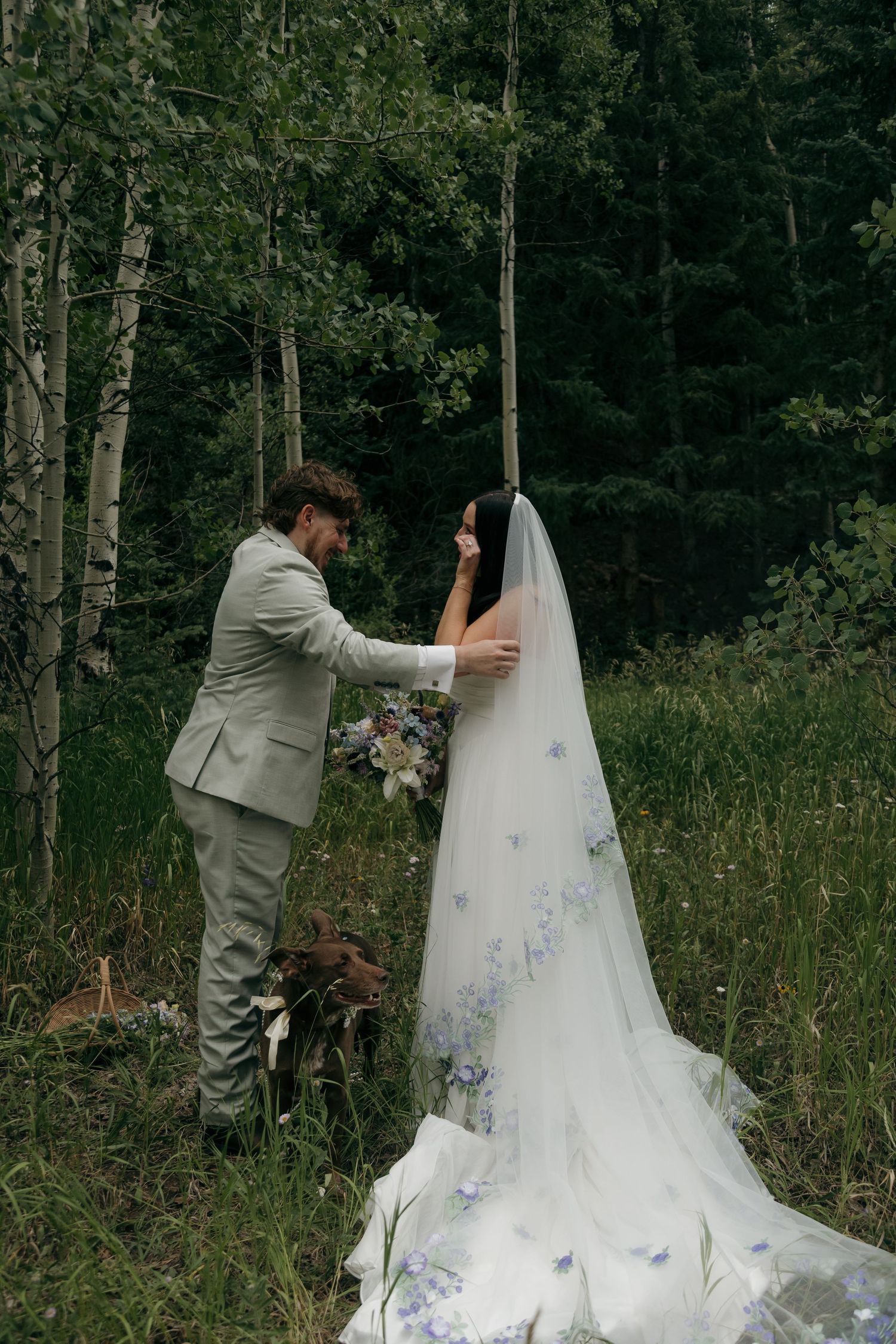 Groom in grey suit tenderly adjusts bride's blue floral cathedral veil in green forest setting.