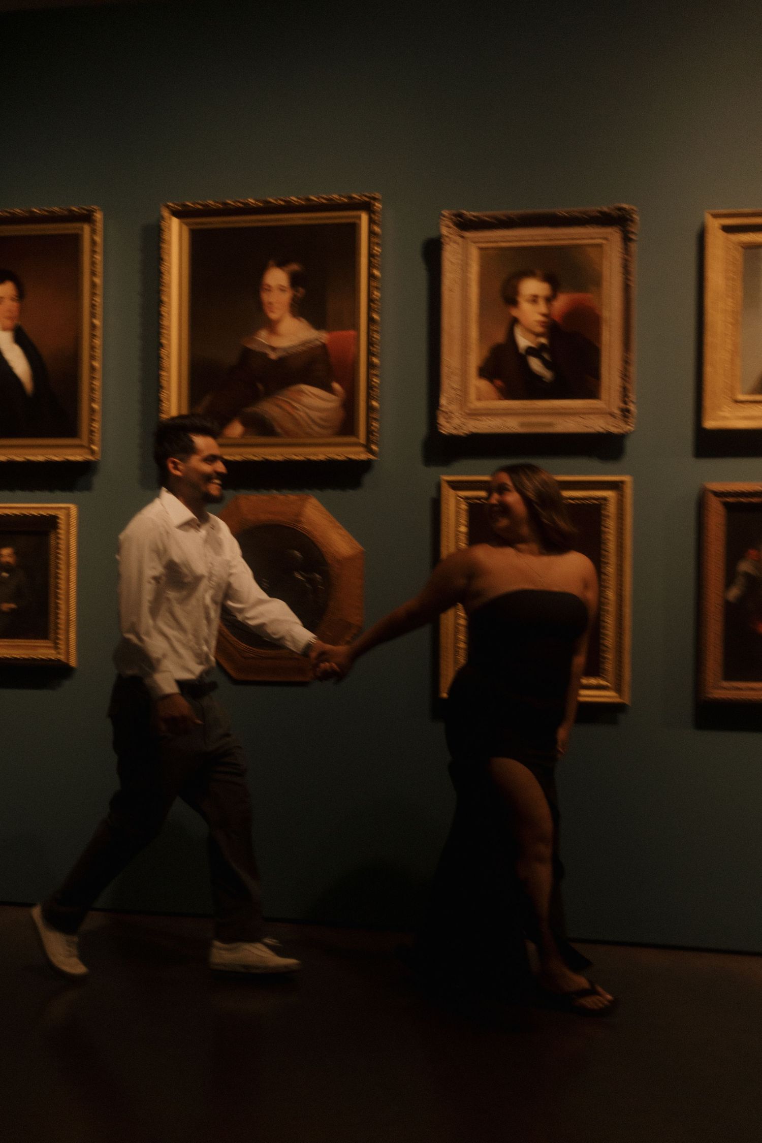 Couple holding hands and dancing in a dimly lit art museum gallery with classic portrait paintings on teal walls.